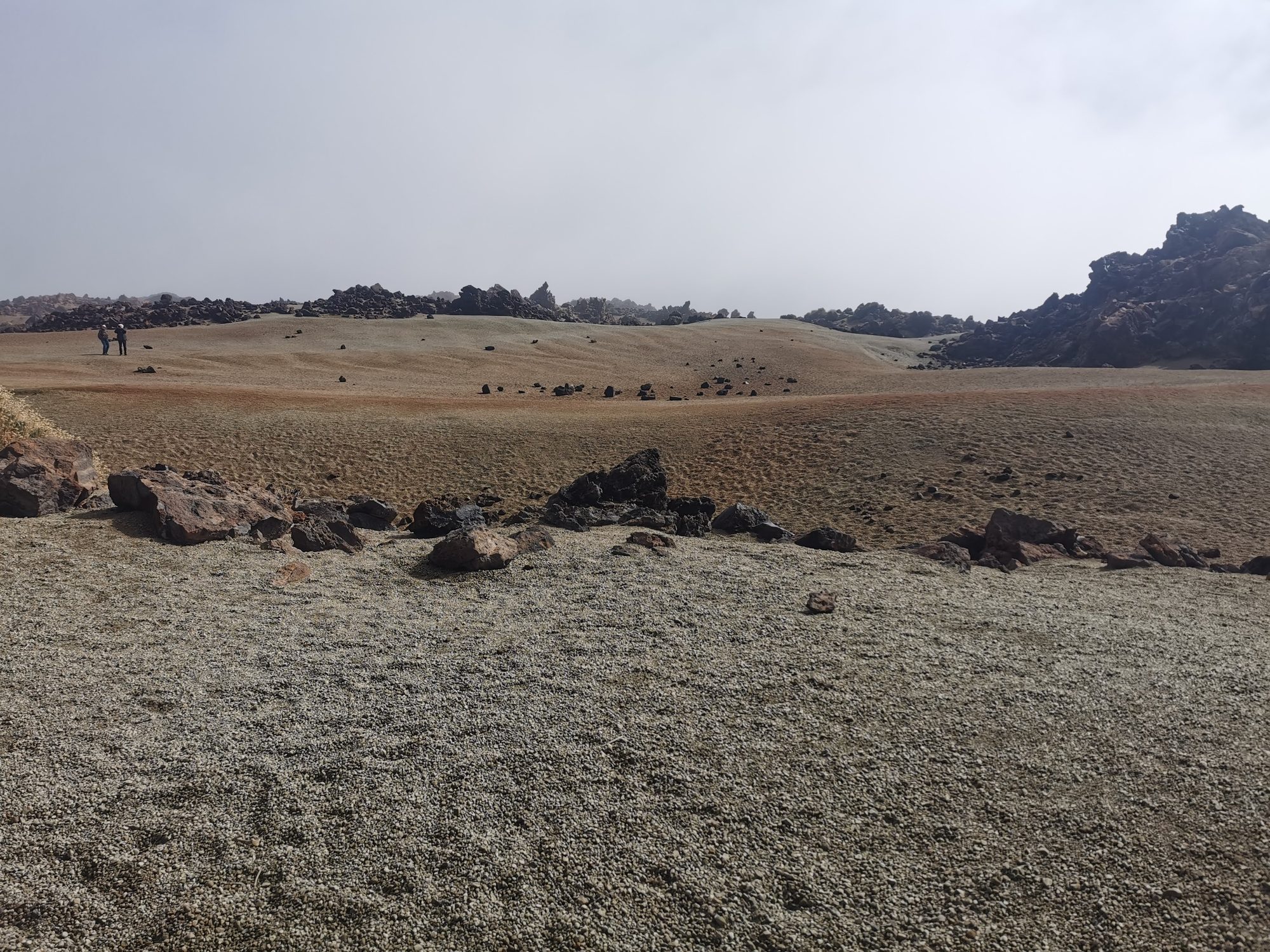 Paisaje volcánico árido con suelo de gravilla y formaciones rocosas oscuras en el horizonte.