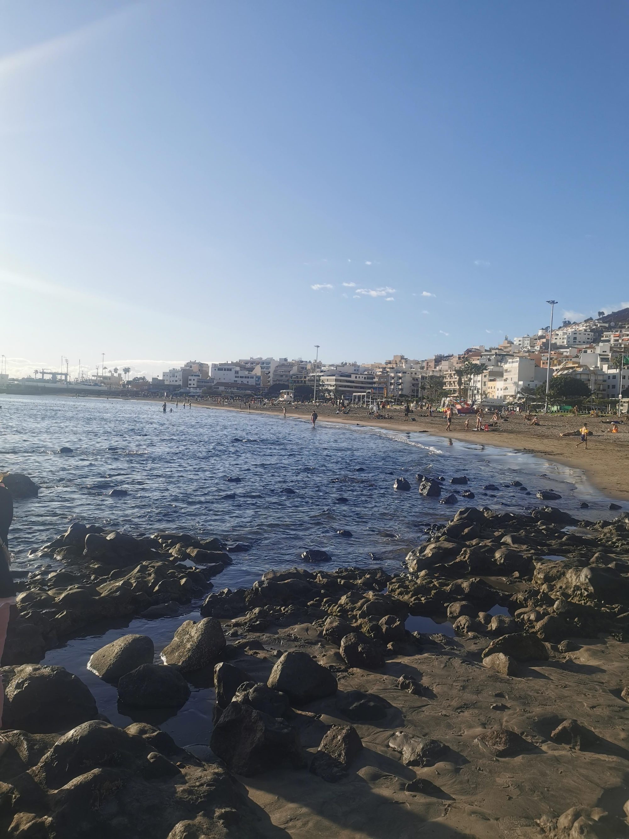 Playa con rocas volcánicas, mar azul y edificios costeros en una tarde soleada.