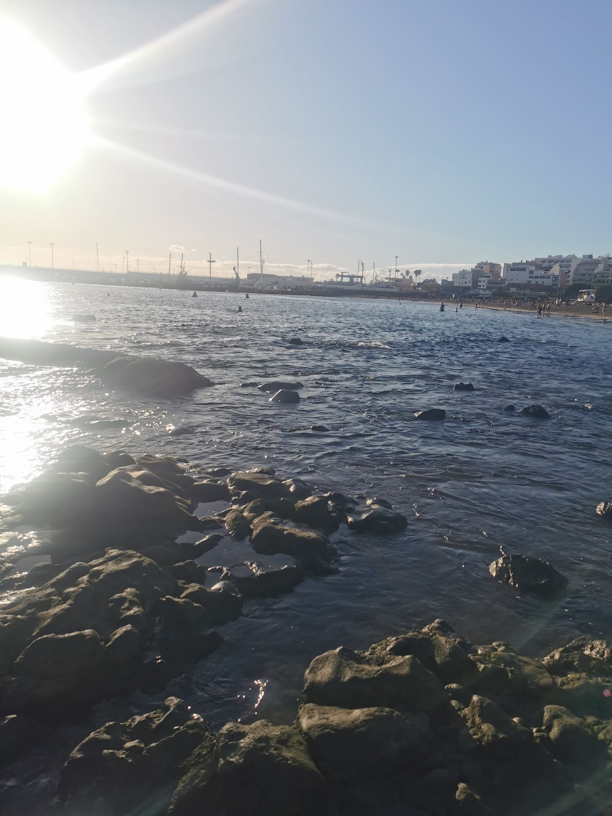 Costa rocosa junto al mar con la silueta urbana al fondo bajo el sol.