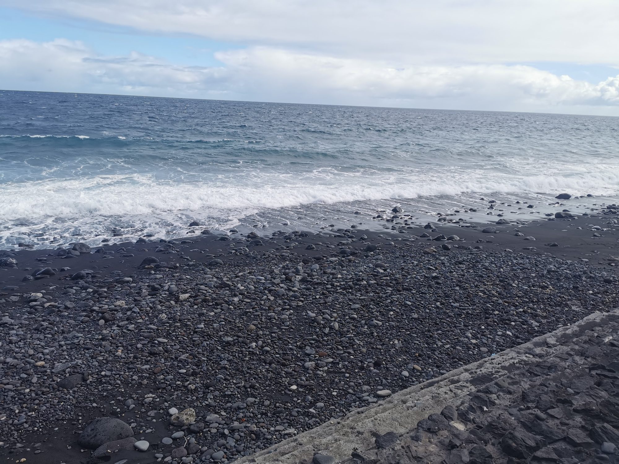Playa de arena negra y piedras volcánicas con olas rompiendo frente al mar abierto.