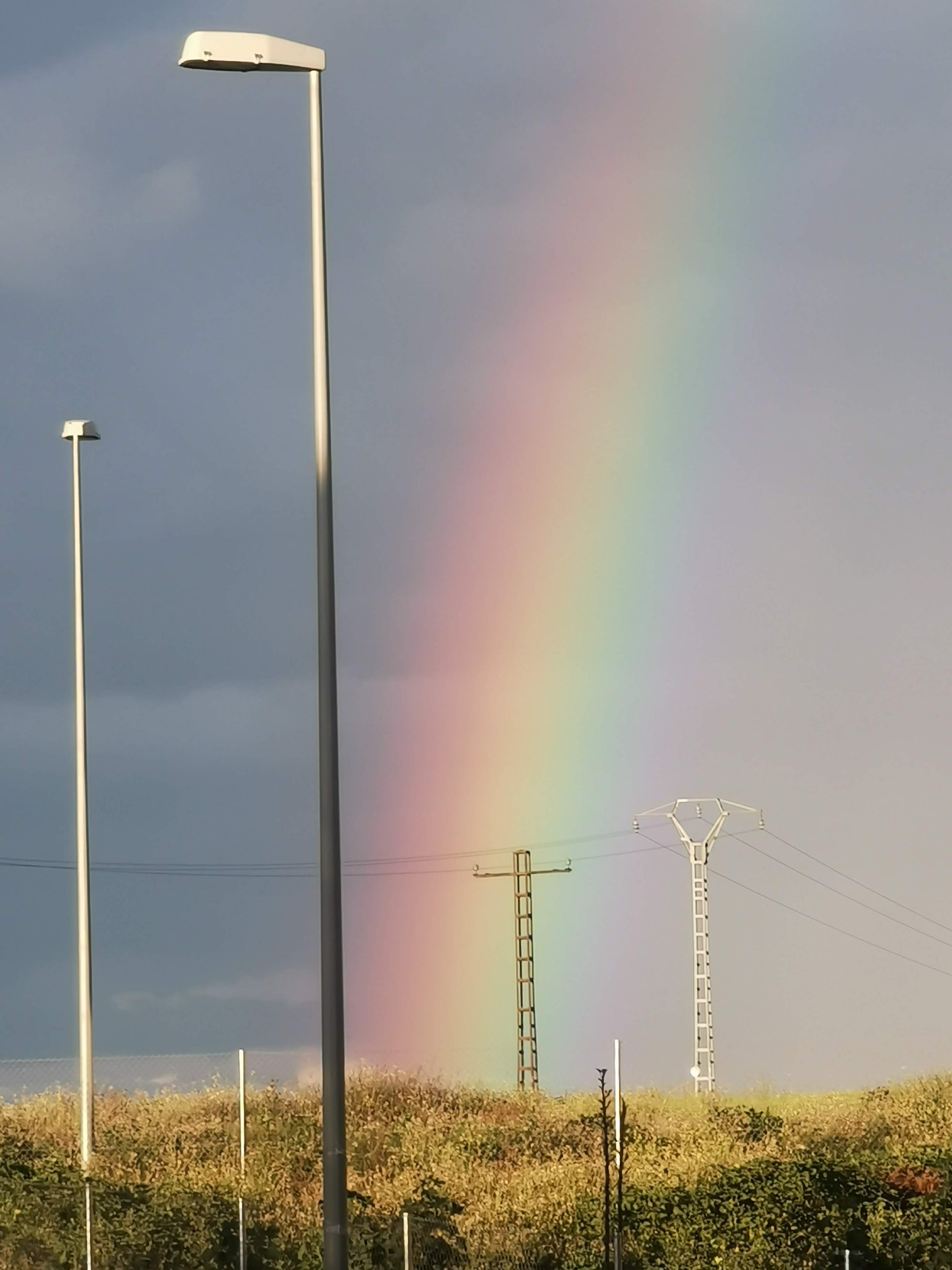 Arcoíris brillante en el cielo tras farolas y torres eléctricas en el campo.
