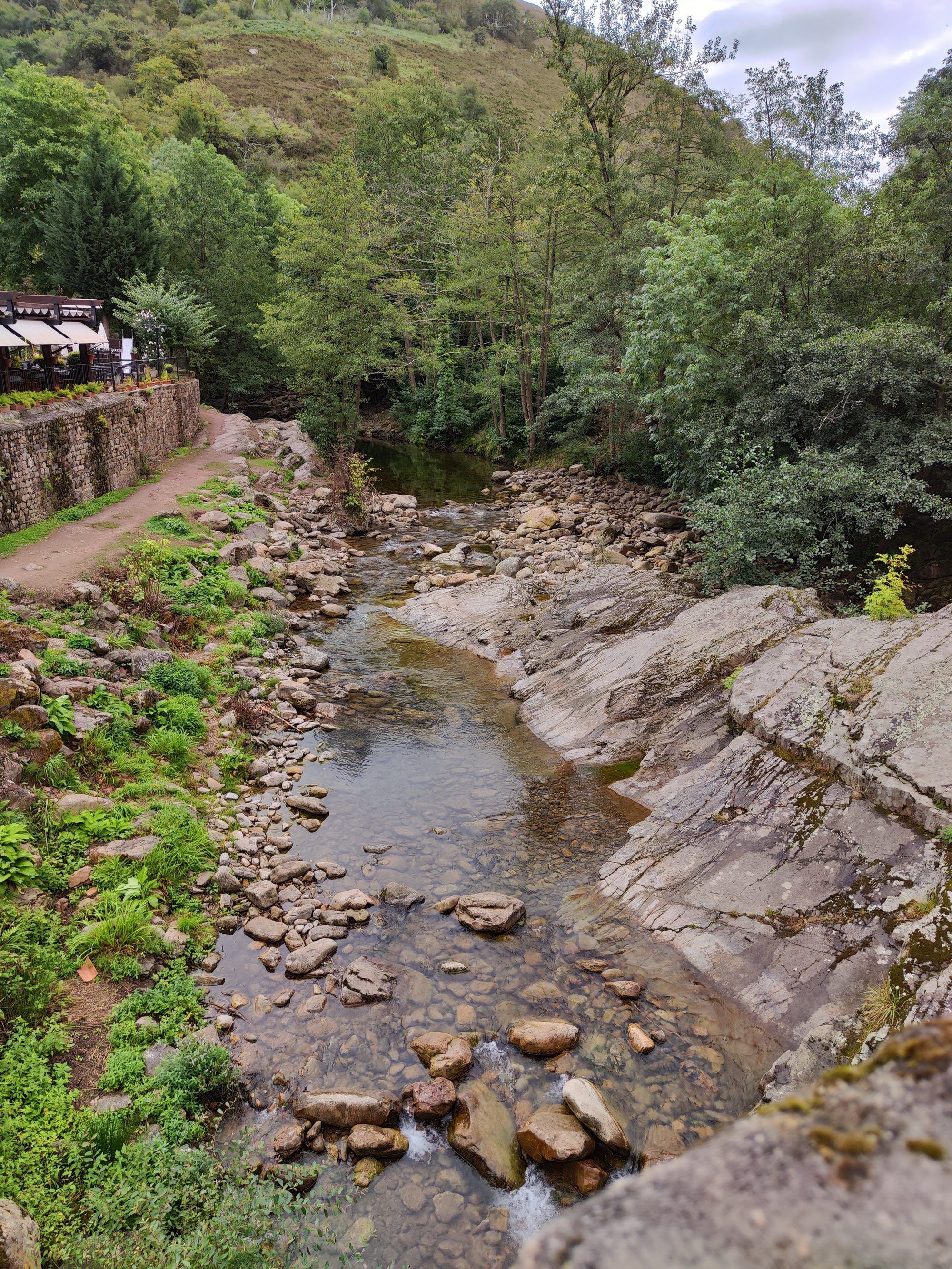 Río pedregoso fluyendo entre frondosa vegetación y laderas montañosas junto a un sendero.