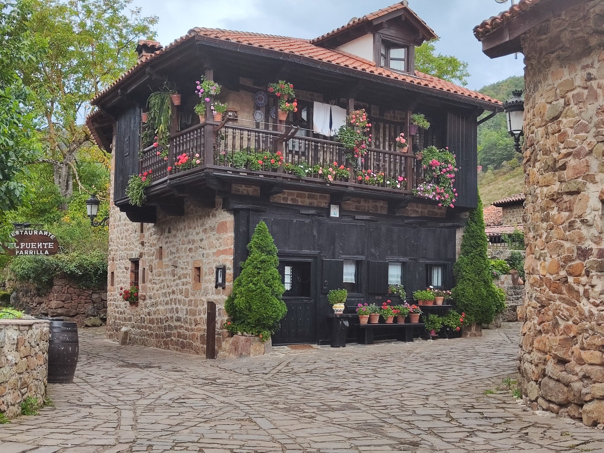 Casa tradicional de piedra y madera con balcón lleno de flores en calle empedrada.