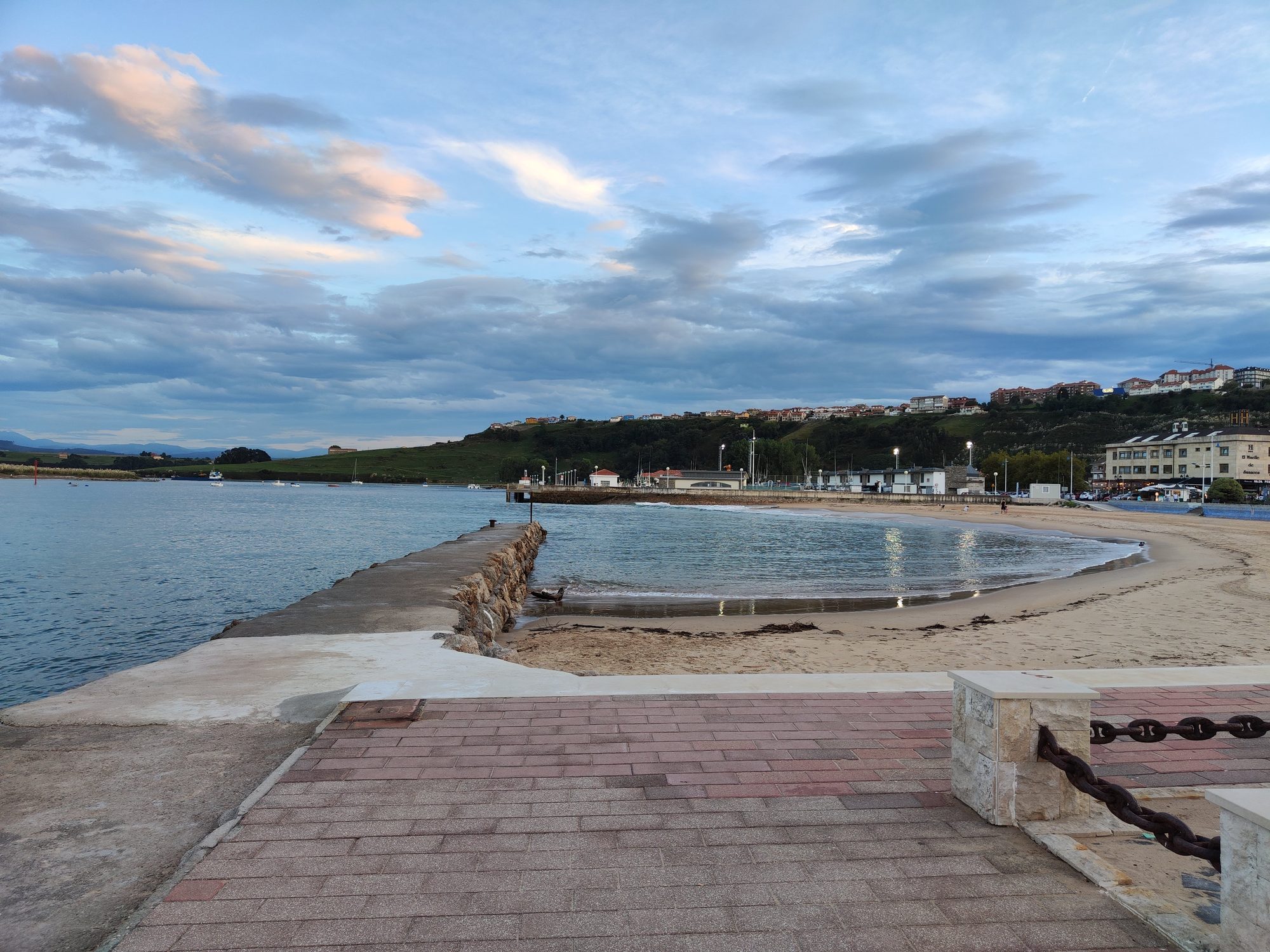 Paseo marítimo, espigón de piedra y playa al atardecer en Suances frente a colinas.