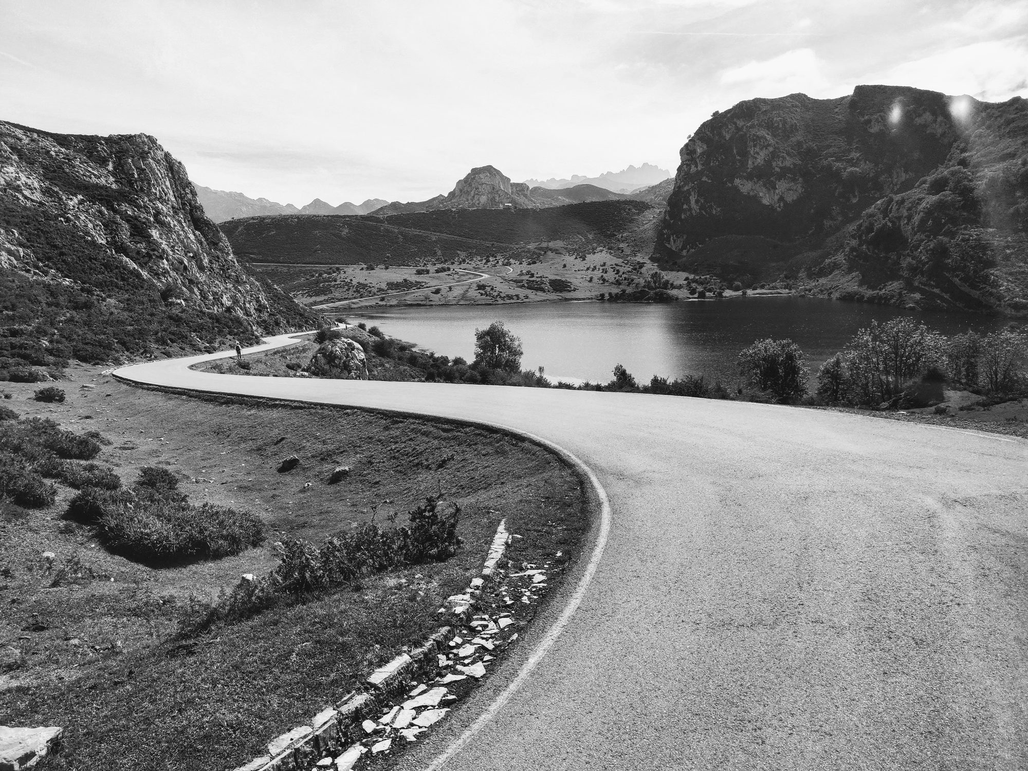 Carretera sinuosa en blanco y negro junto a un lago de montaña entre colinas.
