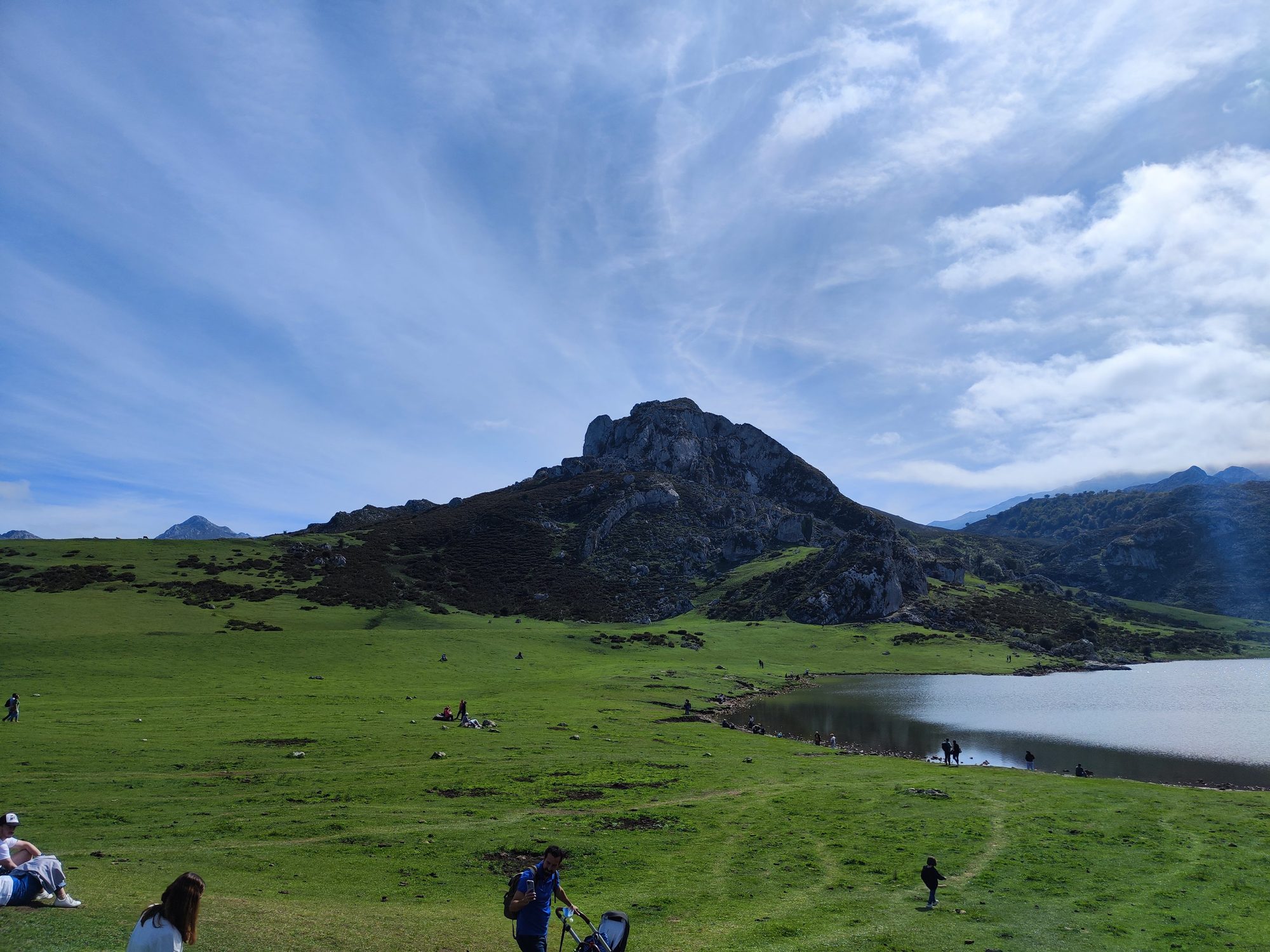 Pradera verde junto a un lago de montaña bajo un cielo azul con nubes.