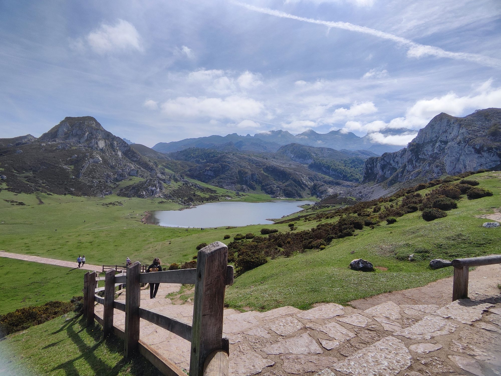 Sendero de piedra hacia un lago de montaña entre verdes colinas y picos rocosos.