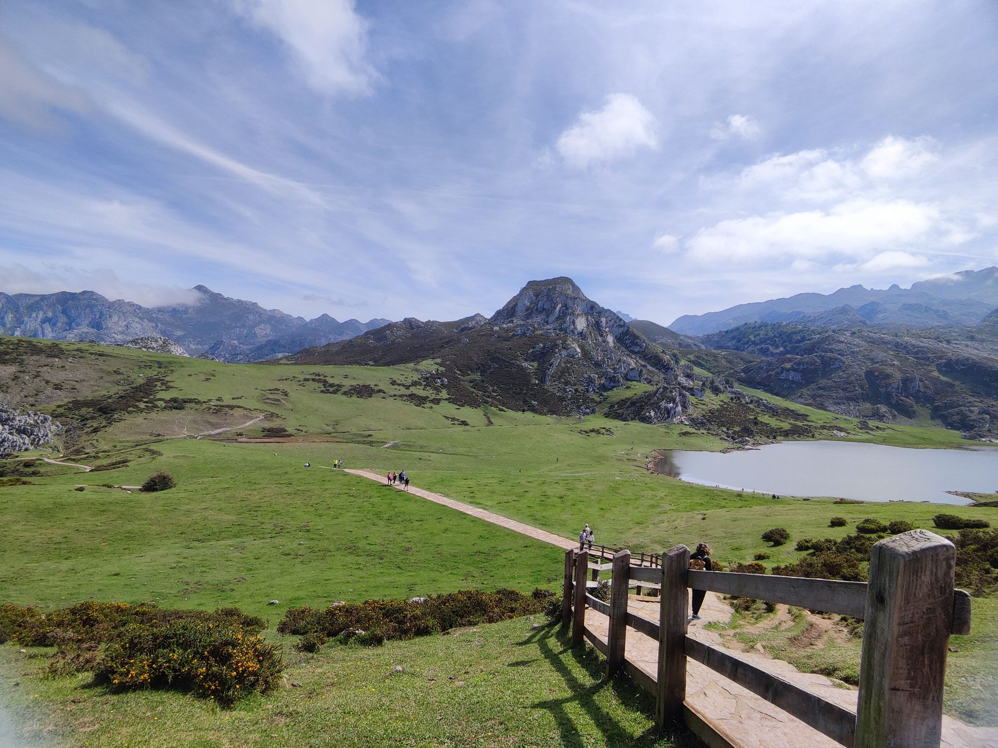 Sendero de madera hacia praderas verdes y lago rodeado de montañas escarpadas.