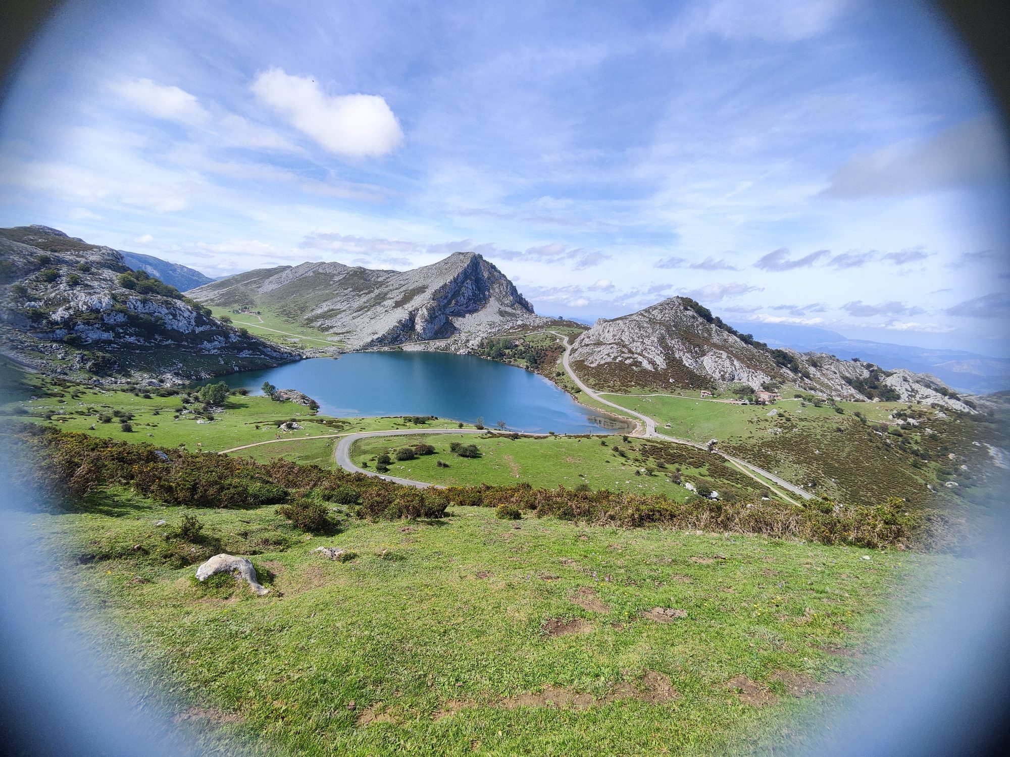 Lago de montaña rodeado de verdes praderas y picos rocosos bajo el cielo azul.