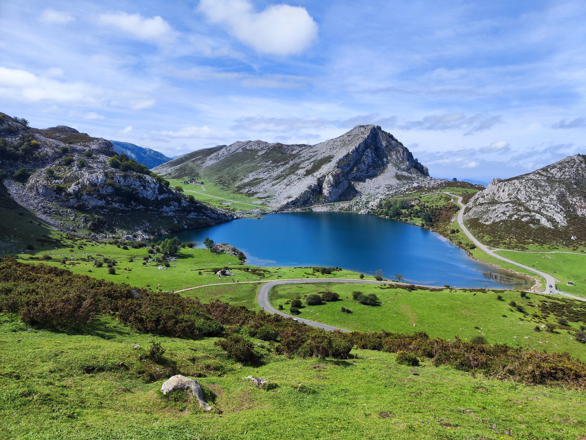 Lago de alta montaña rodeado de verdes praderas y picos rocosos bajo cielo azul.