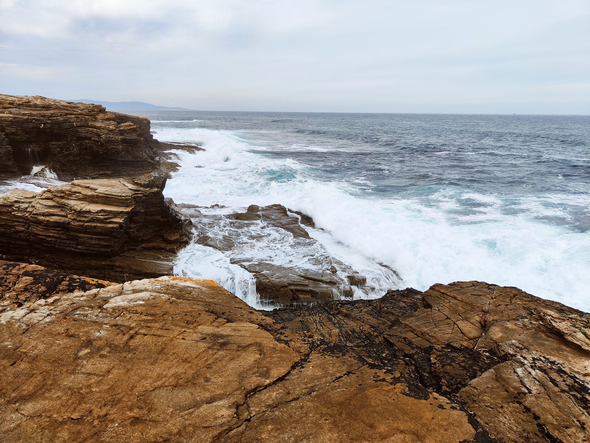 Olas del mar Cantábrico rompiendo con fuerza contra acantilados rocosos bajo cielo nublado.