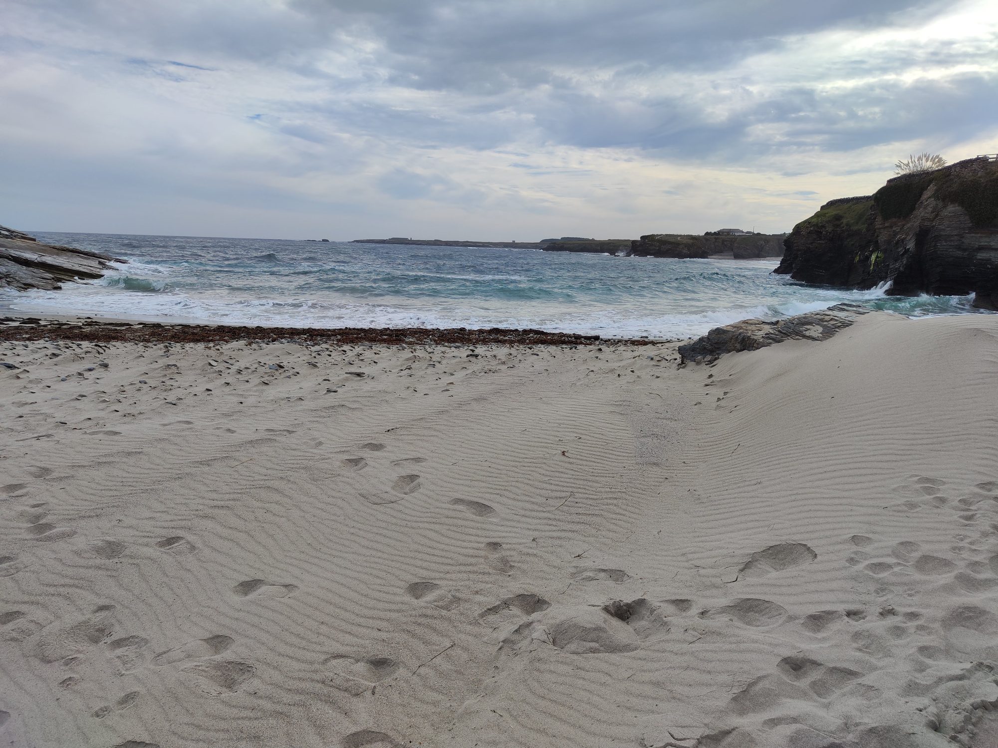 Playa de arena fina con dunas y olas entre acantilados rocosos bajo cielo nublado.