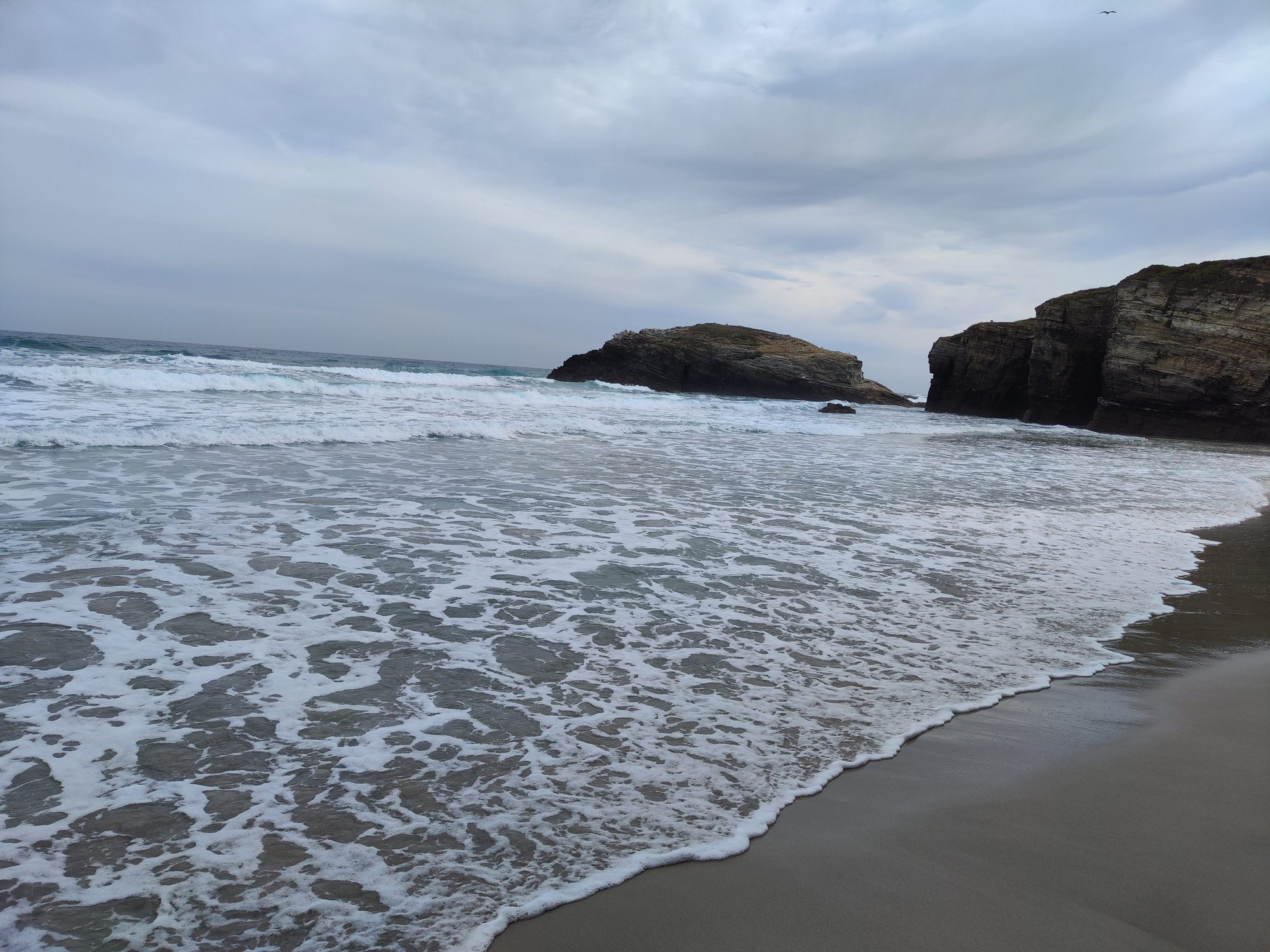 Olas rompiendo en la orilla de una playa con acantilados bajo un cielo nublado.
