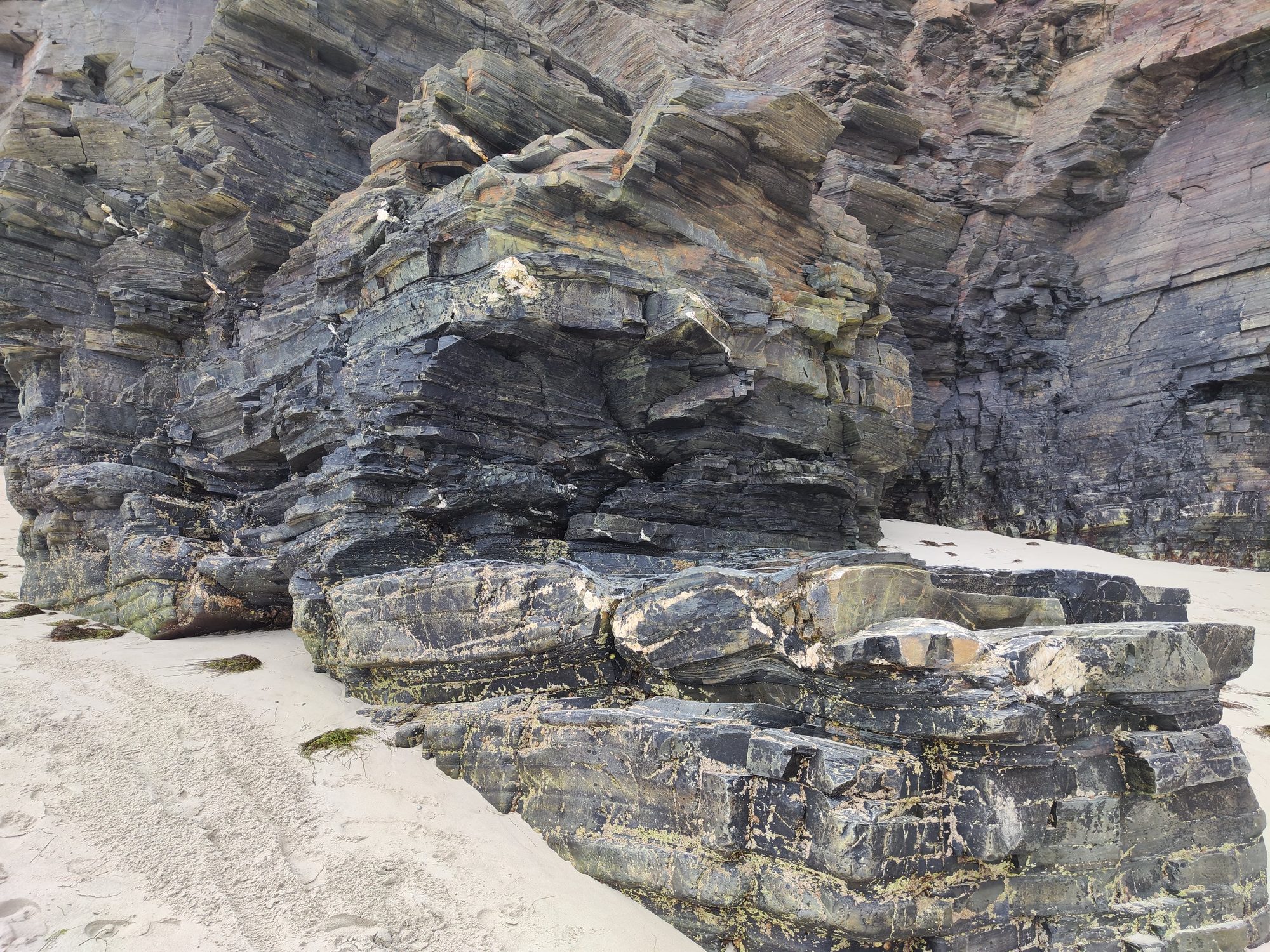 Grandes formaciones rocosas de pizarra estratificada sobre la arena fina de una playa.
