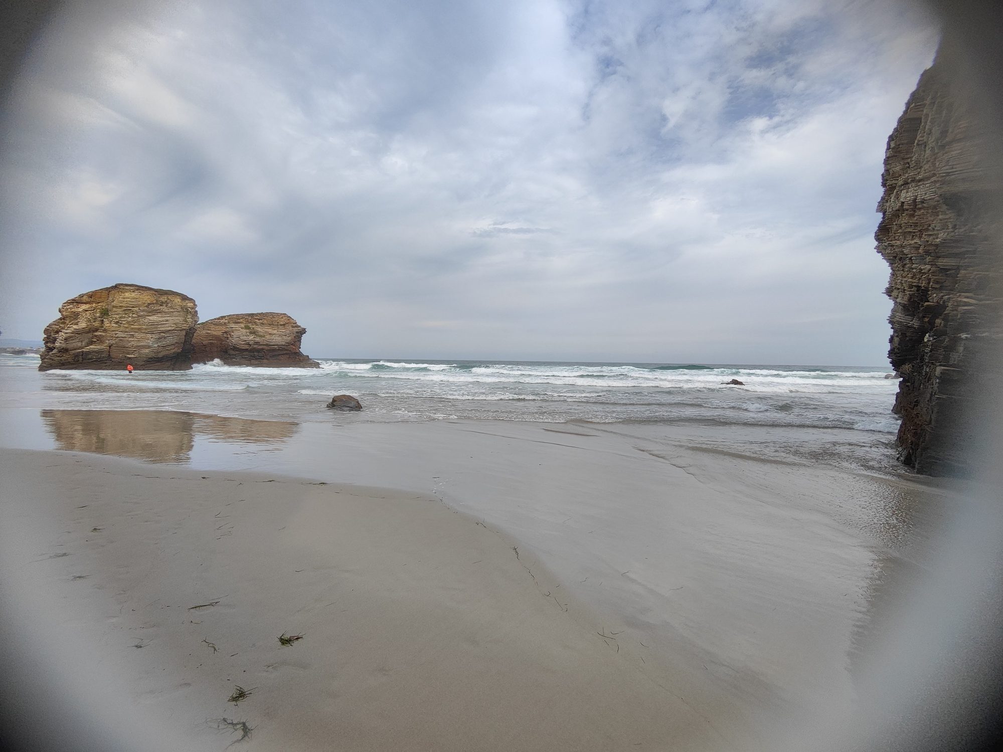 Formaciones rocosas y olas sobre la arena húmeda de la playa con cielo nublado.