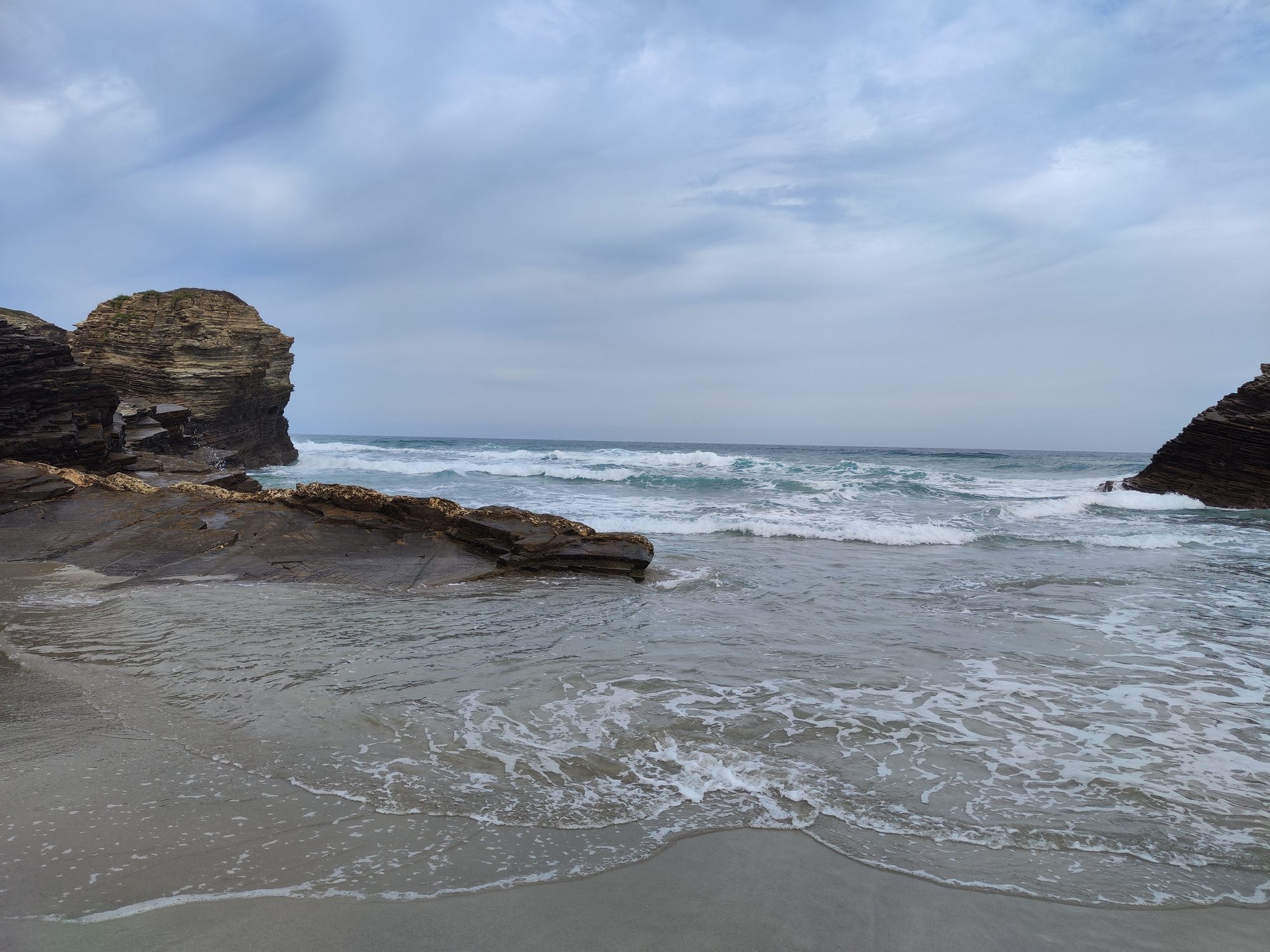 Acantilados rocosos y olas rompiendo en la orilla arenosa bajo un cielo nublado.