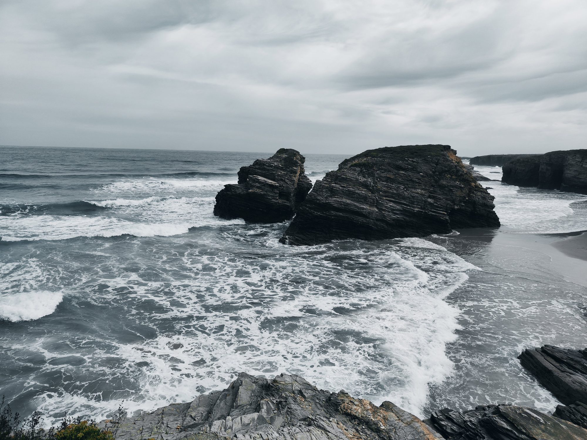 Grandes formaciones rocosas en el mar con fuerte oleaje bajo un cielo nublado.