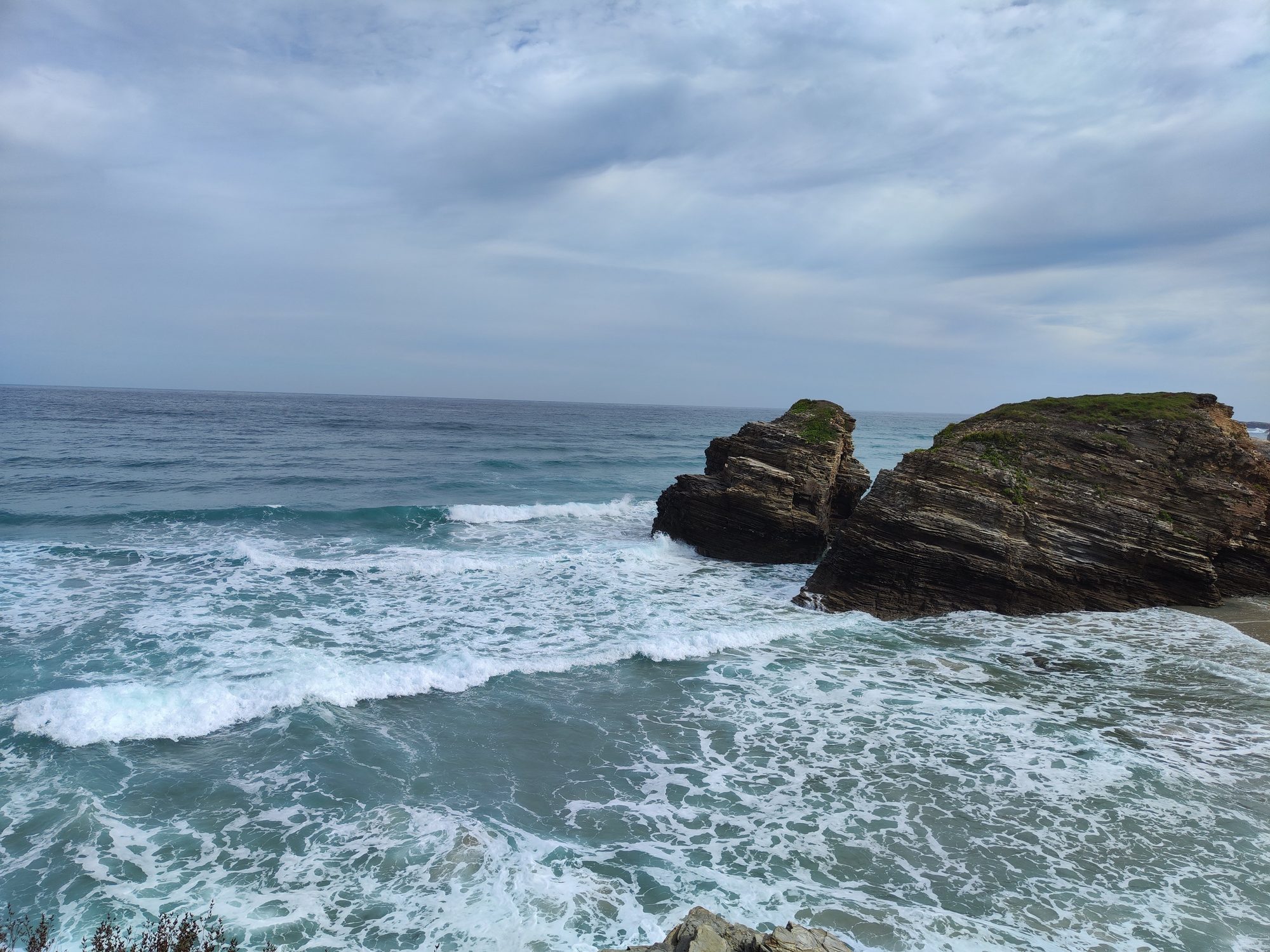 Olas del mar rompiendo contra formaciones rocosas en la costa bajo cielo nublado.