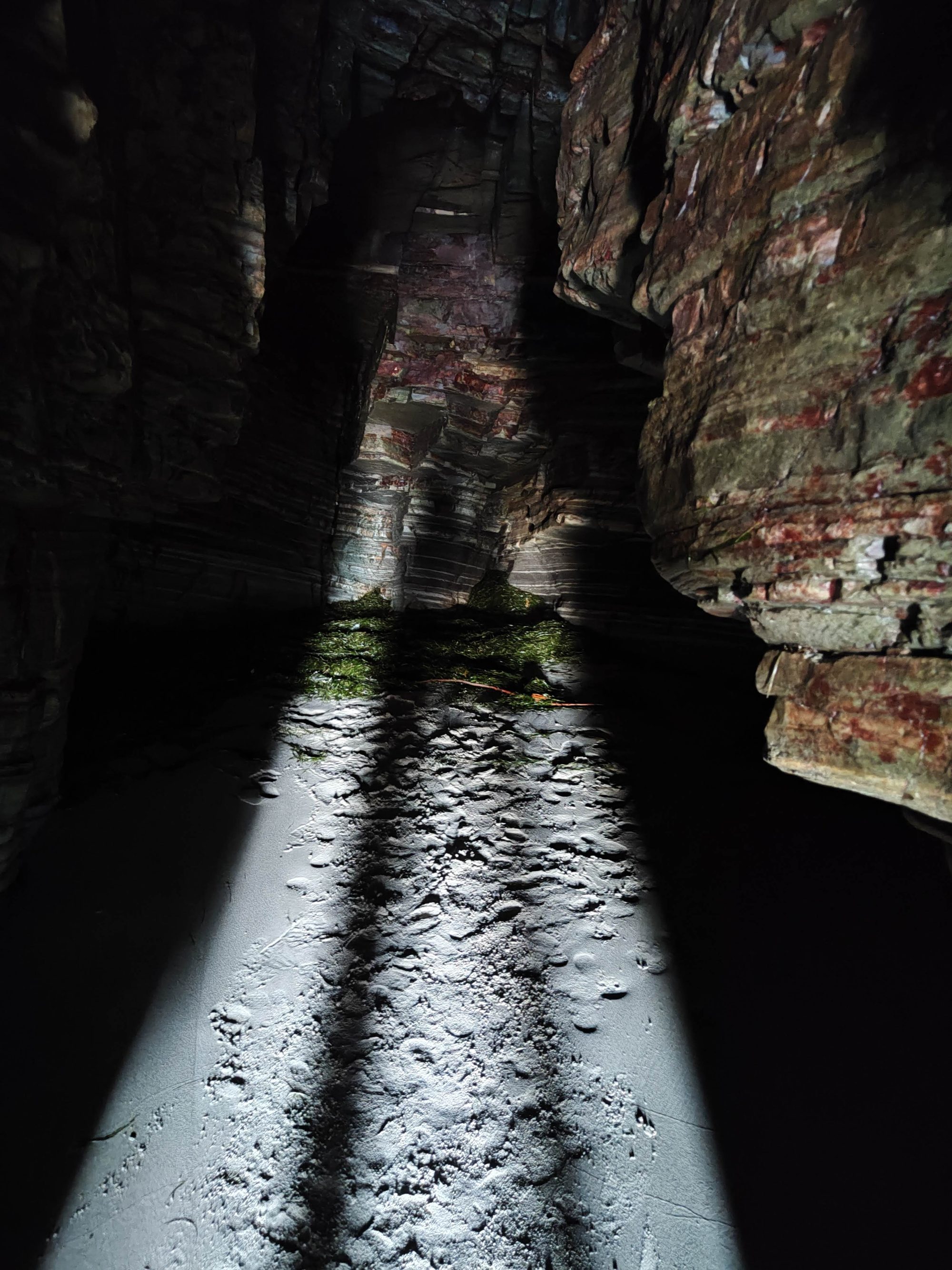 Interior de una cueva marina con paredes de roca estratificada y suelo arenoso iluminado.