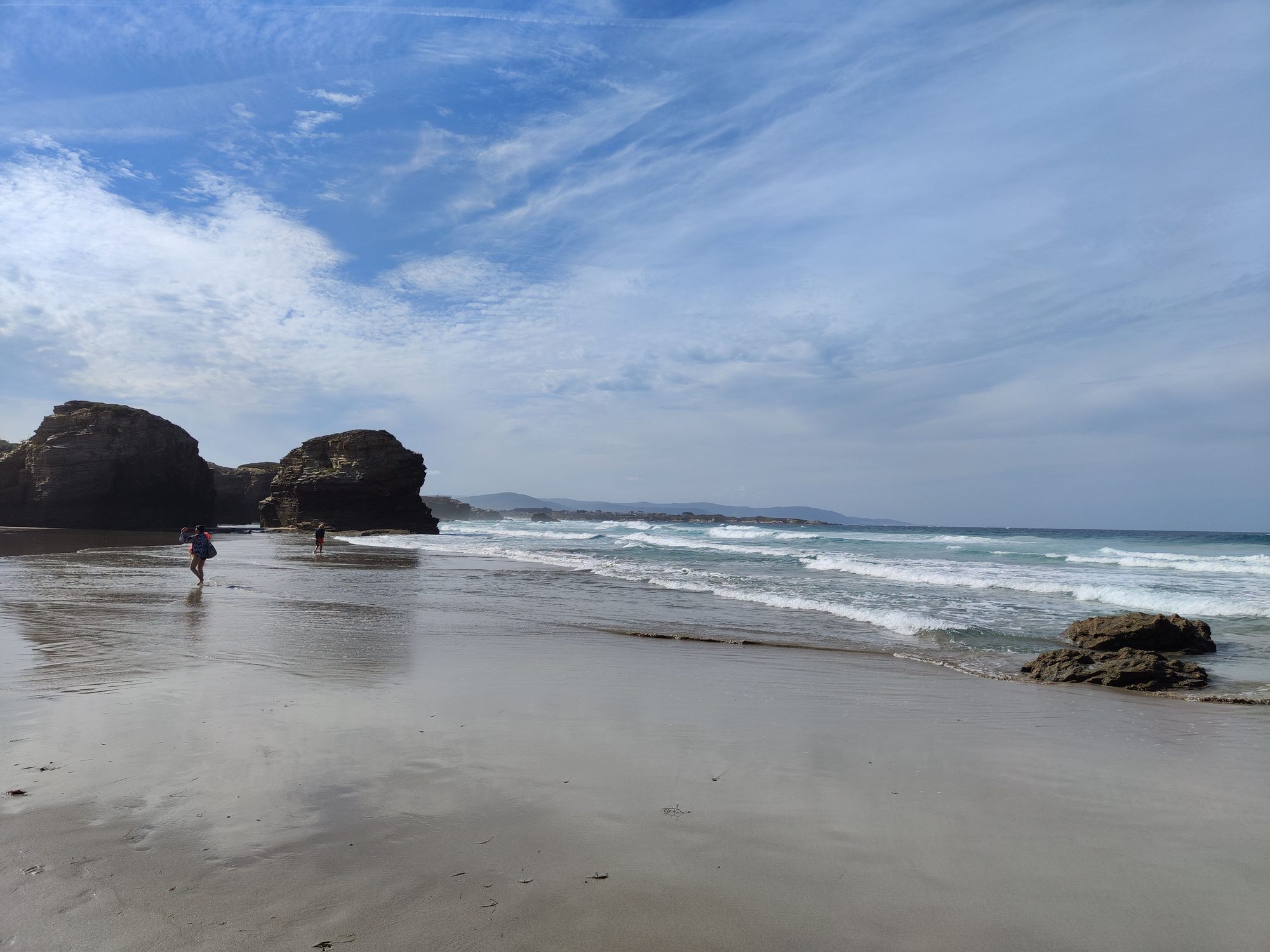 Playa amplia de arena mojada con formaciones rocosas y olas bajo un cielo azul.