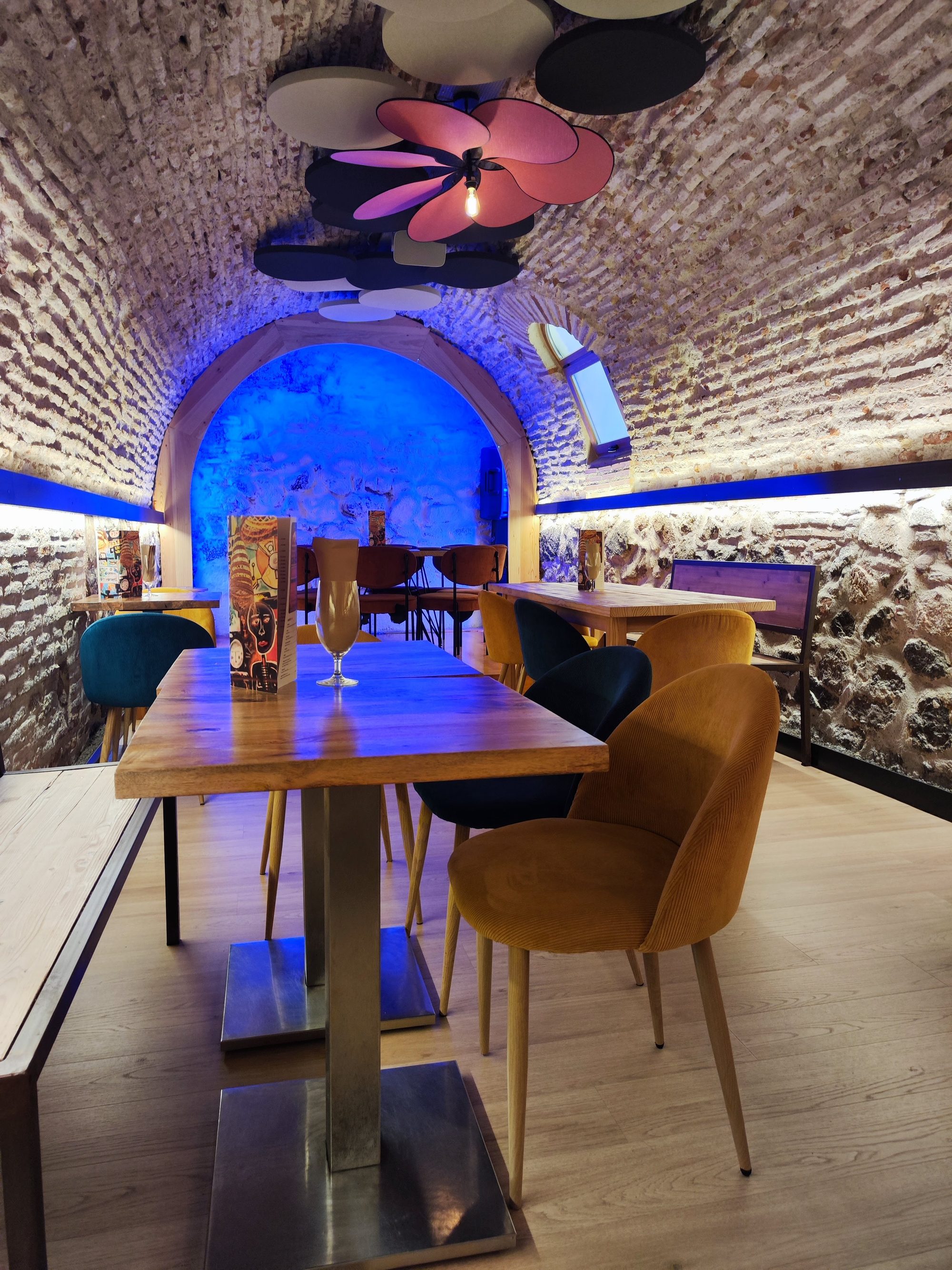 Bóveda de ladrillo en restaurante con iluminación azul, mesas de madera y sillas de colores.