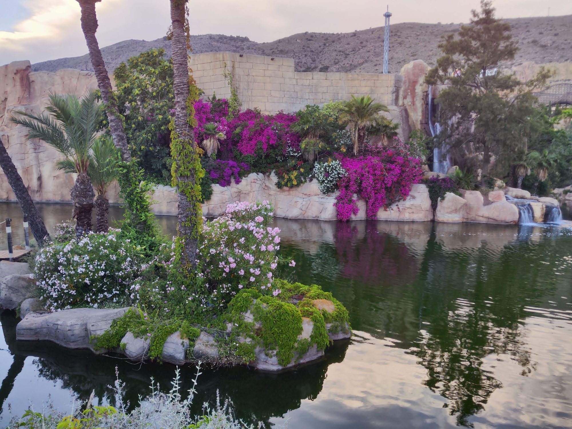Lago rodeado de exuberante vegetación, buganvillas en flor y cascadas sobre paredes de piedra.