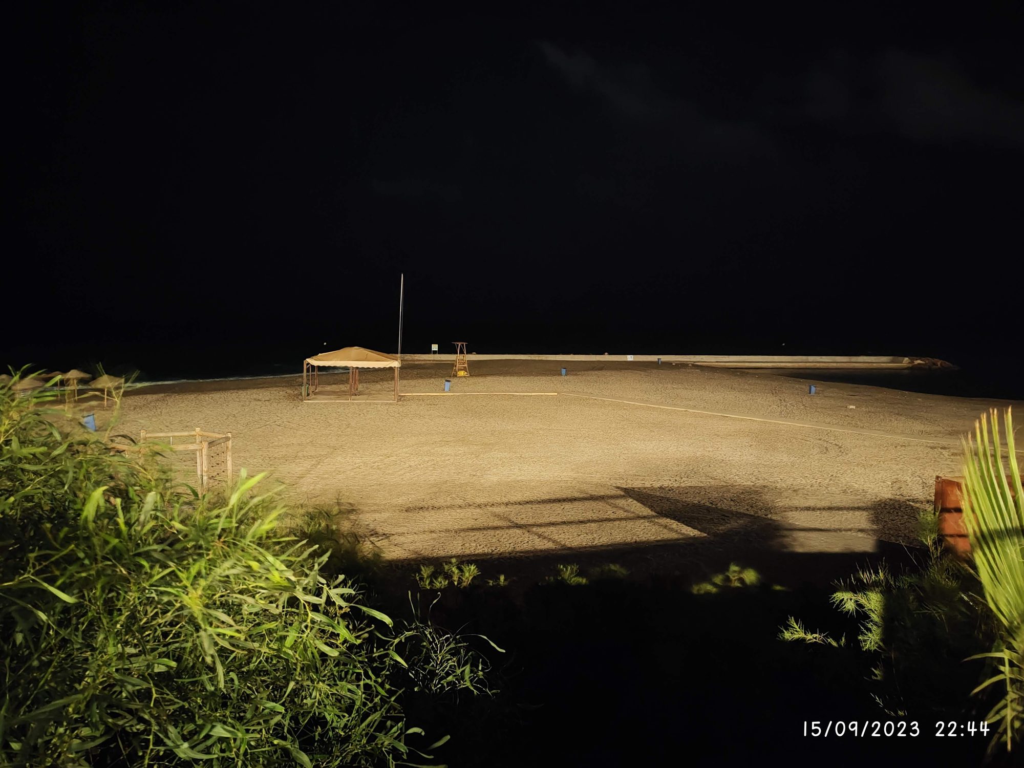 Playa iluminada de noche con pérgola de madera, torre de socorrista y vegetación frontal.