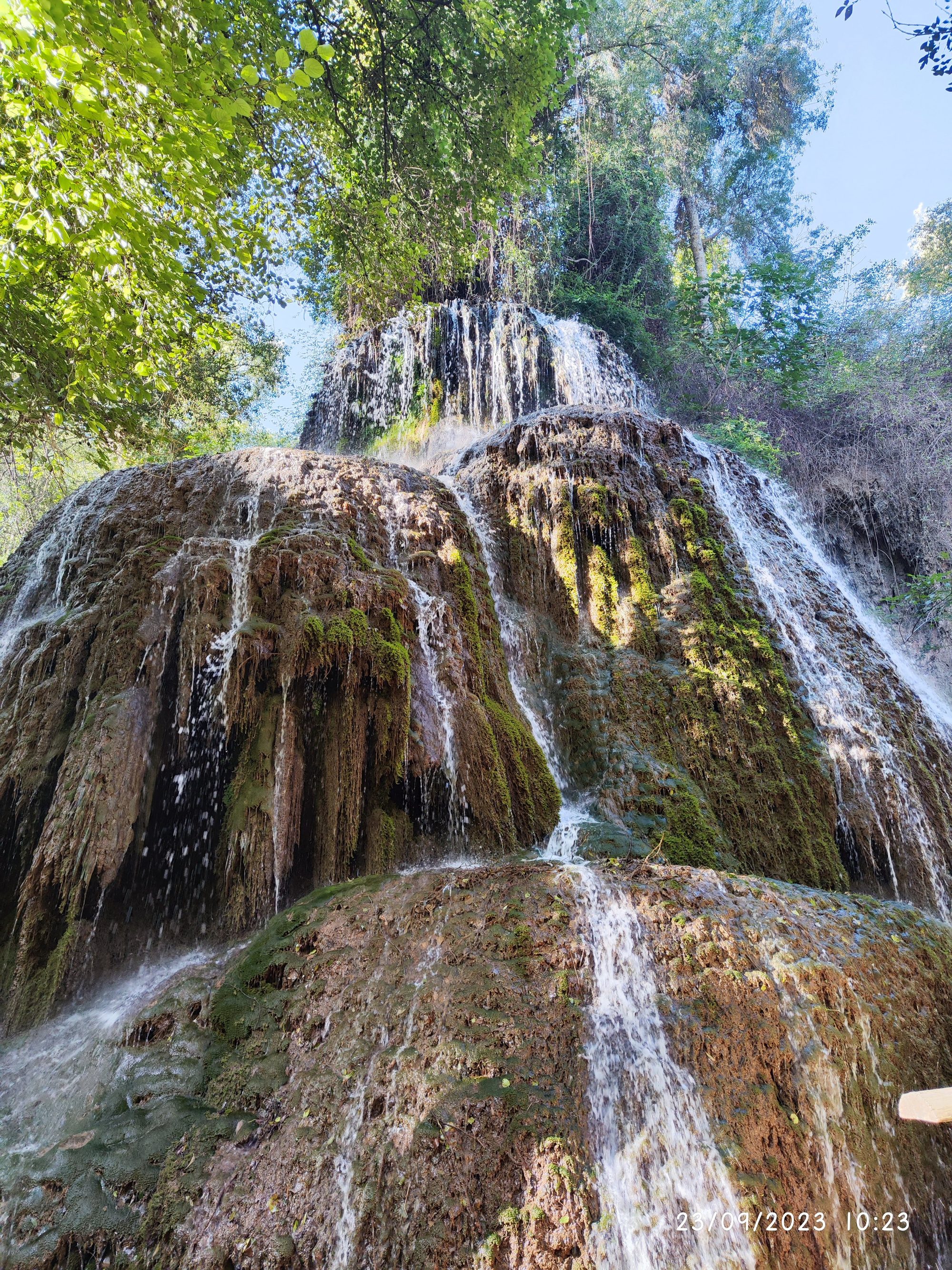 Gran cascada de agua fluyendo sobre rocas cubiertas de musgo entre exuberante vegetación.