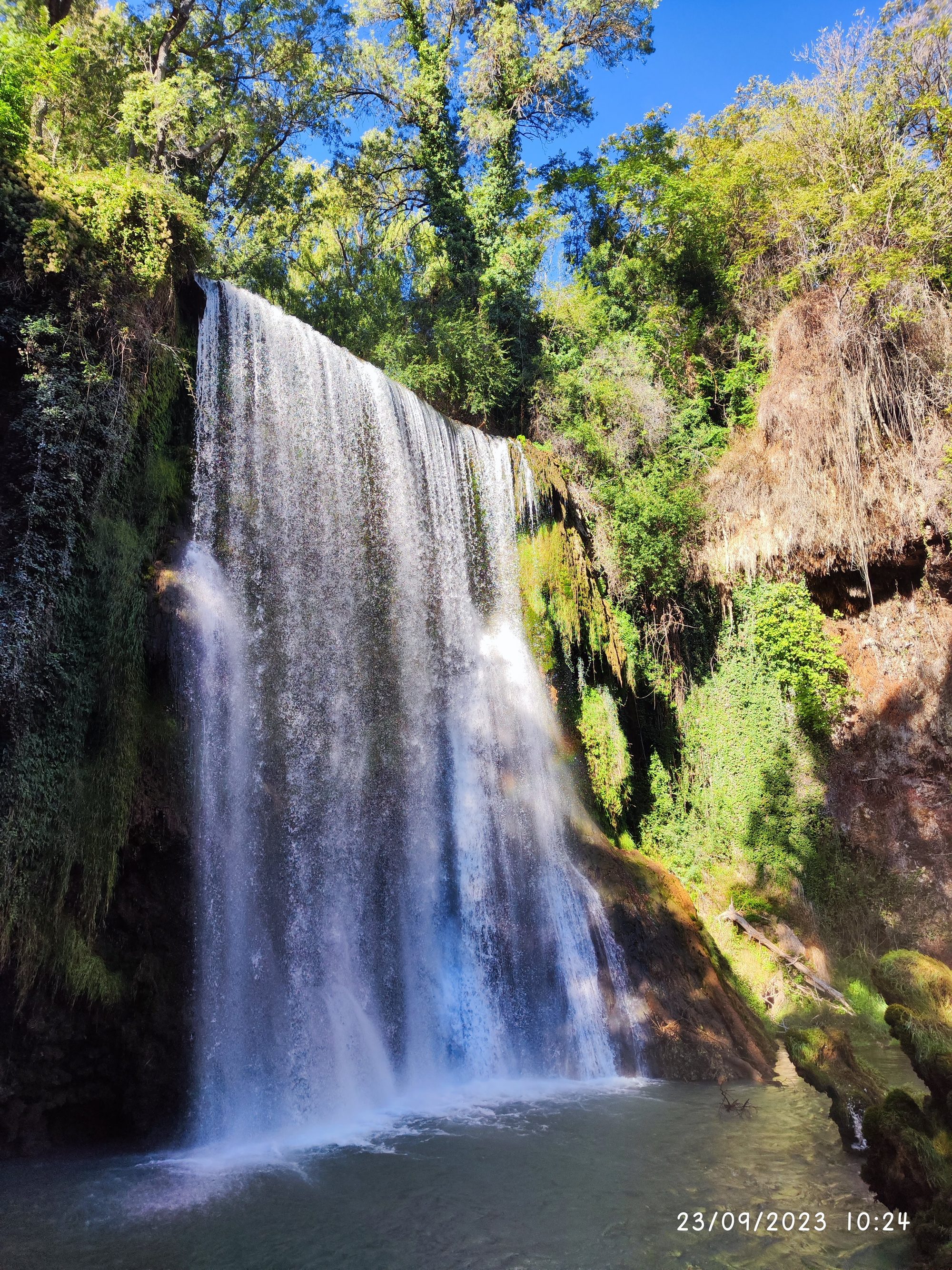 Gran cascada rodeada de frondosa vegetación verde cayendo sobre una poza de agua natural.