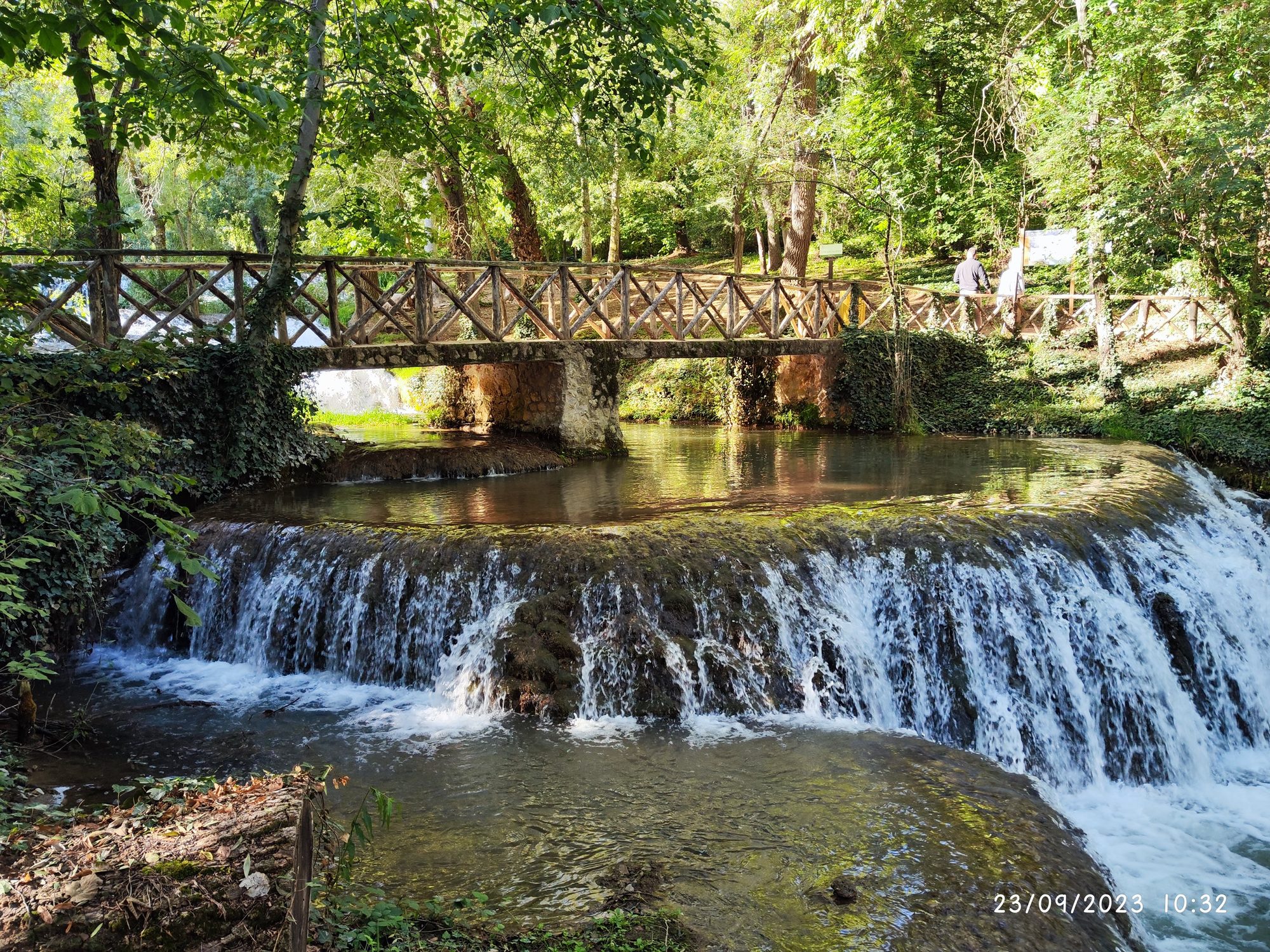 Puente de madera sobre una cascada rodeada de vegetación frondosa en un río.