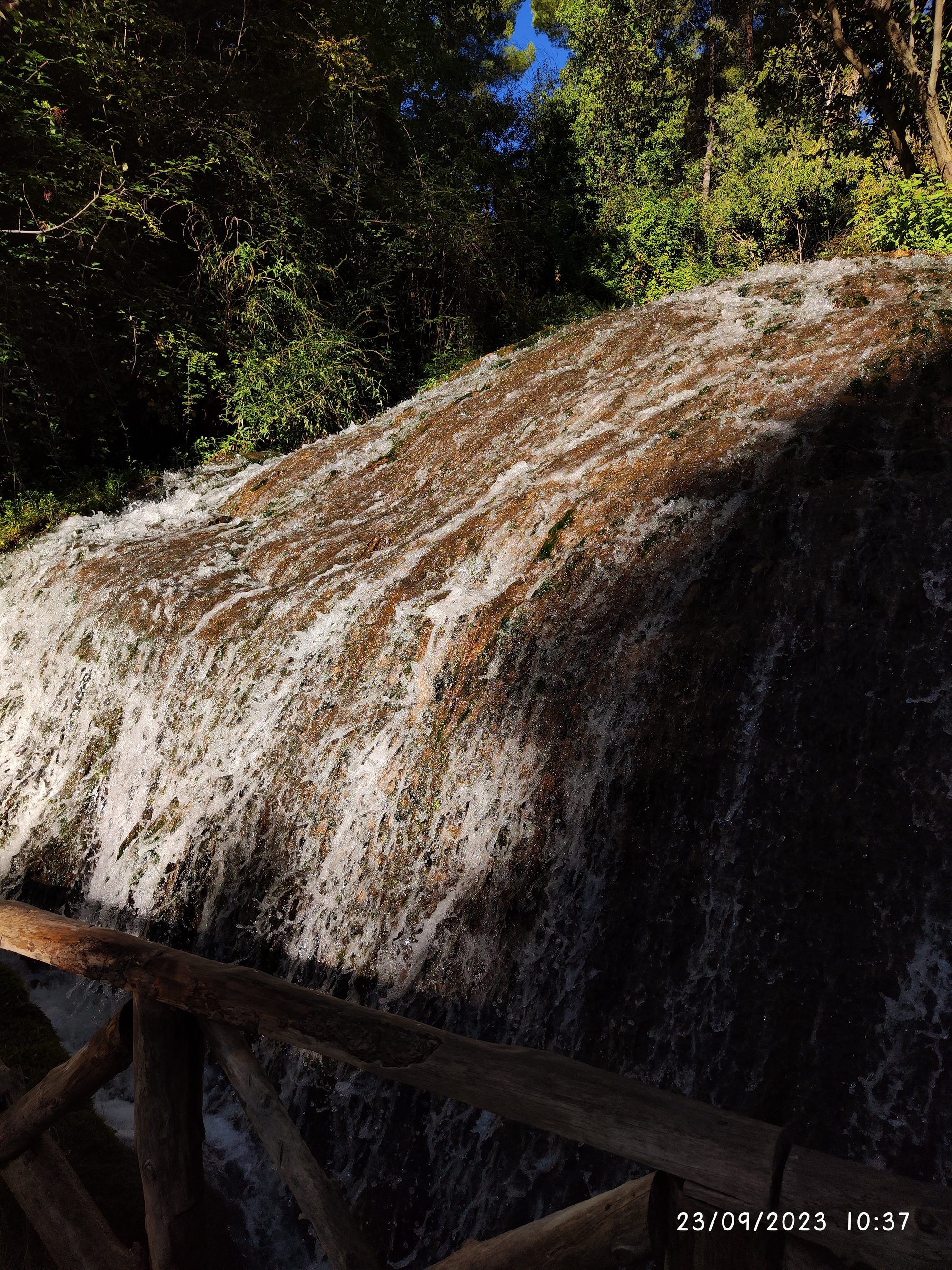Cascada de agua fluyendo sobre rocas rodeada de vegetación verde y barandilla de madera.