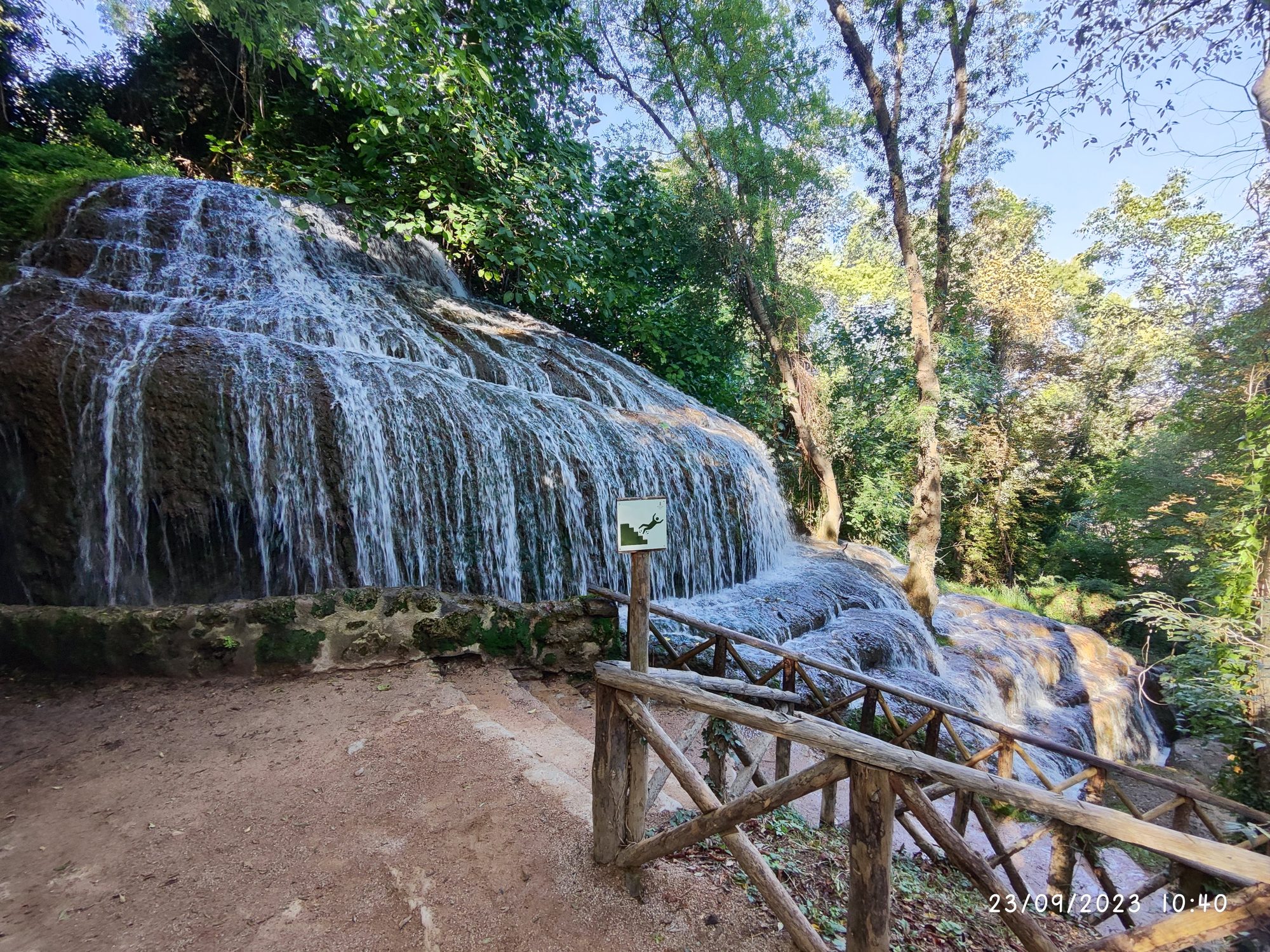 Cascada fluyendo sobre rocas calcáreas en un denso bosque con valla de madera.