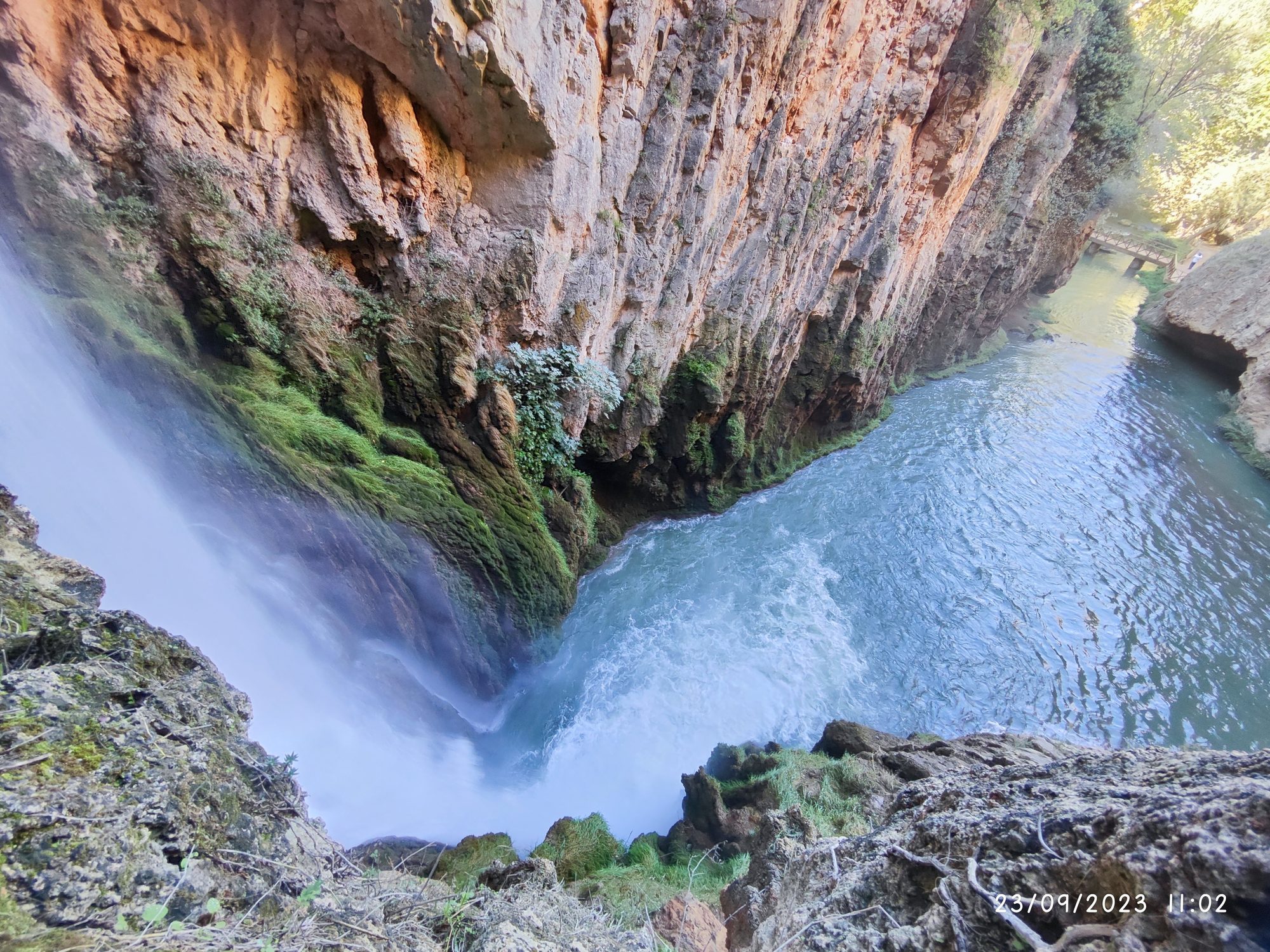Cascada cayendo hacia un río turquesa encajonado entre altas paredes rocosas.