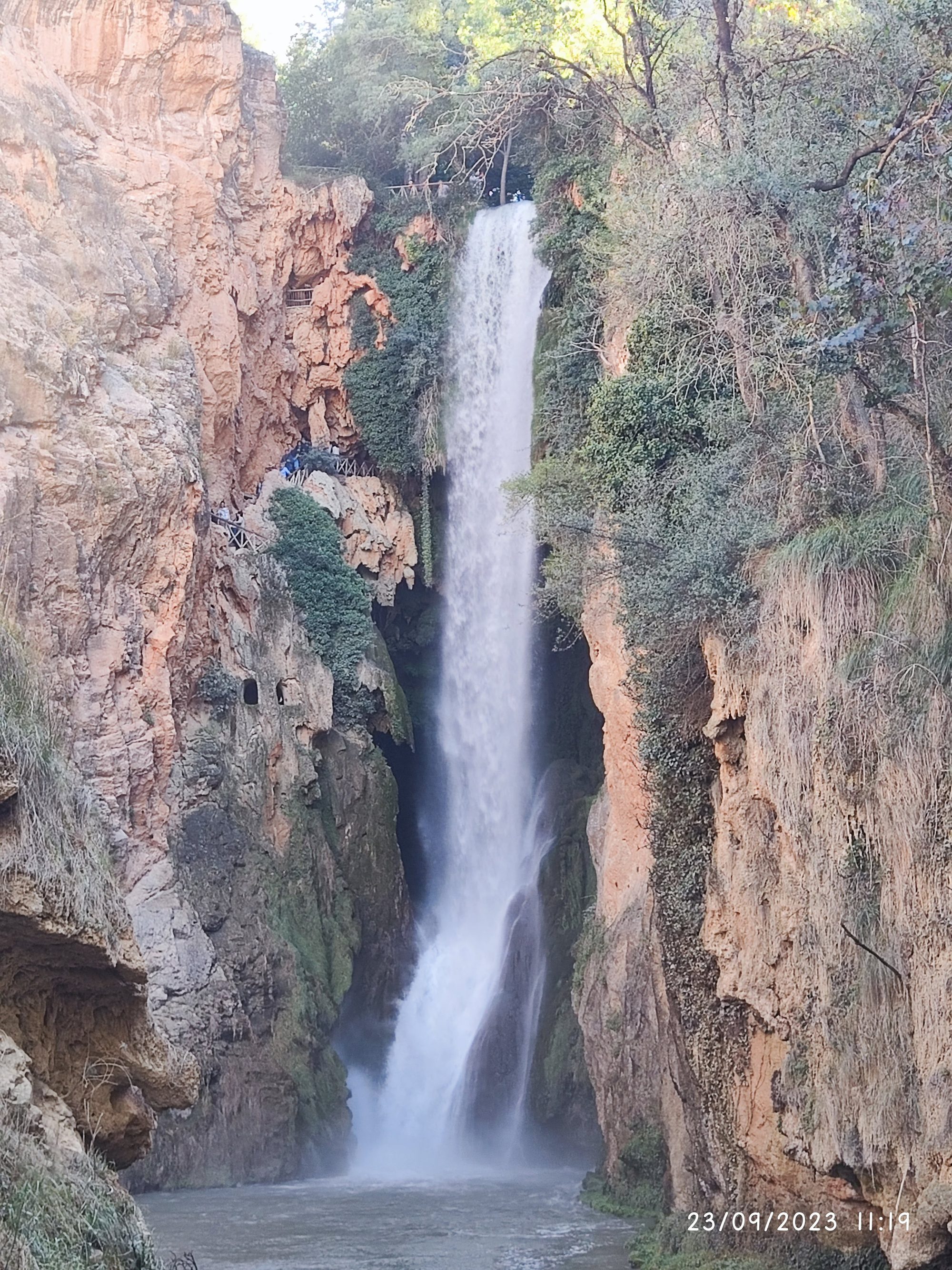 Gran cascada de agua cayendo entre elevados acantilados rocosos y abundante vegetación verde.