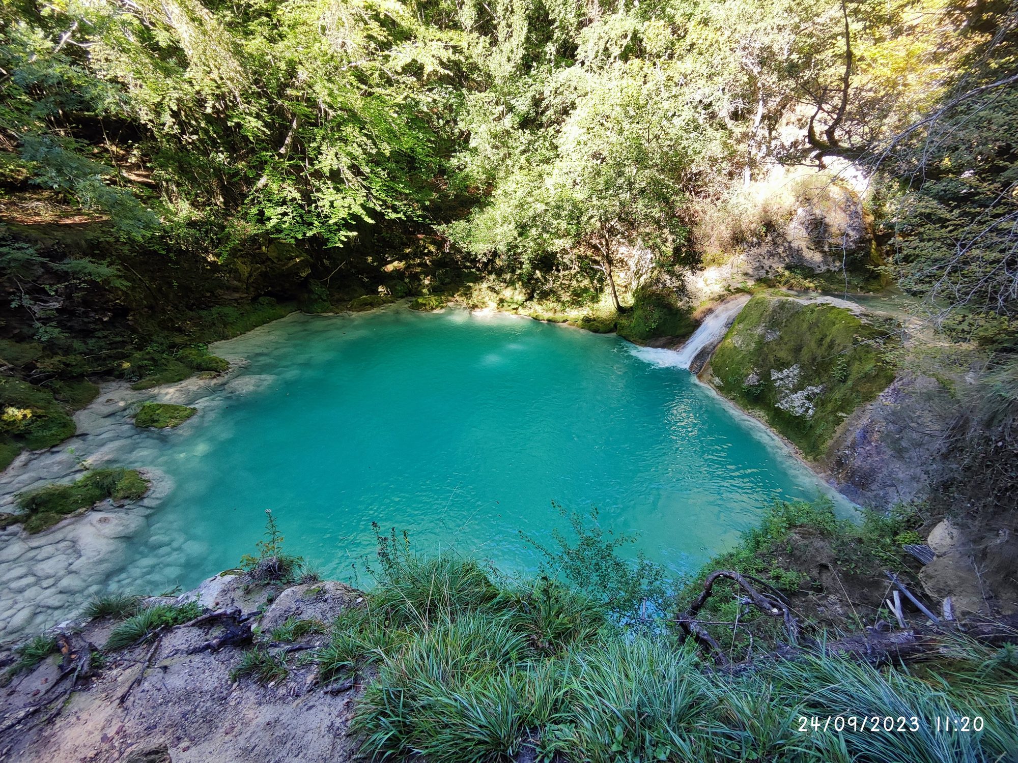 Poza de agua turquesa rodeada de frondoso bosque verde y una pequeña cascada.