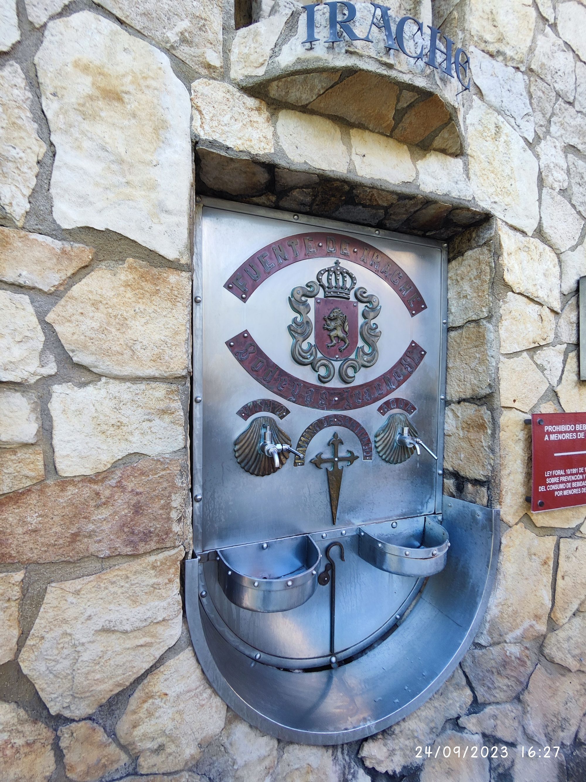 Fuente metálica de vino y agua encastrada en un muro de piedra en Ayegui.
