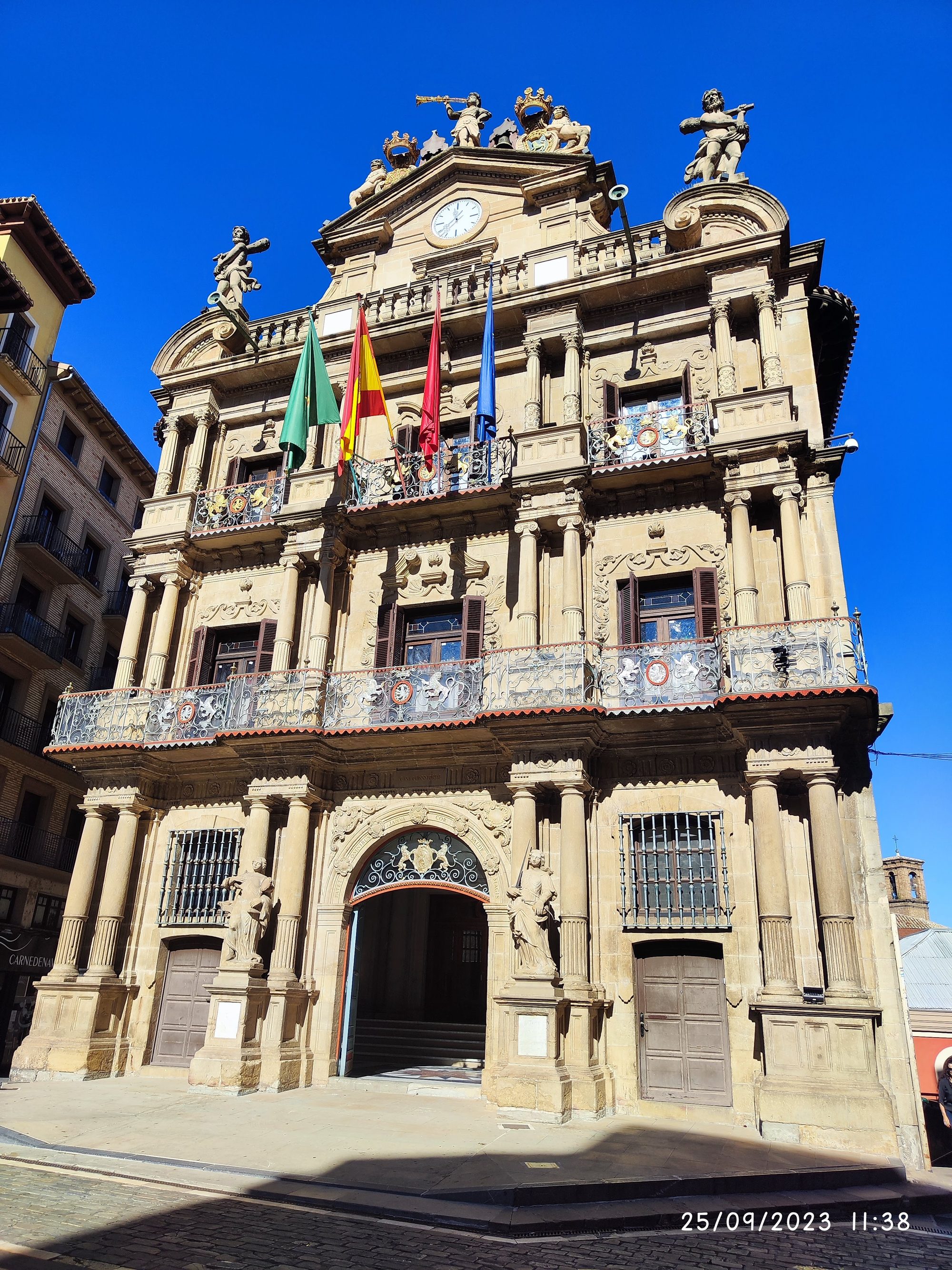 Fachada barroca del Ayuntamiento de Pamplona con banderas y esculturas bajo un cielo azul.