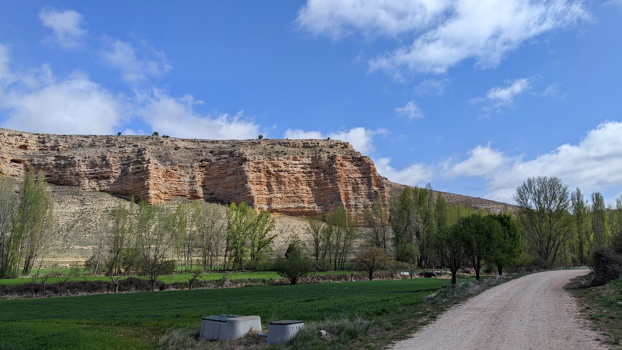 Camino de tierra rodeado de campos verdes frente a un cortado rocoso con cielo azul.