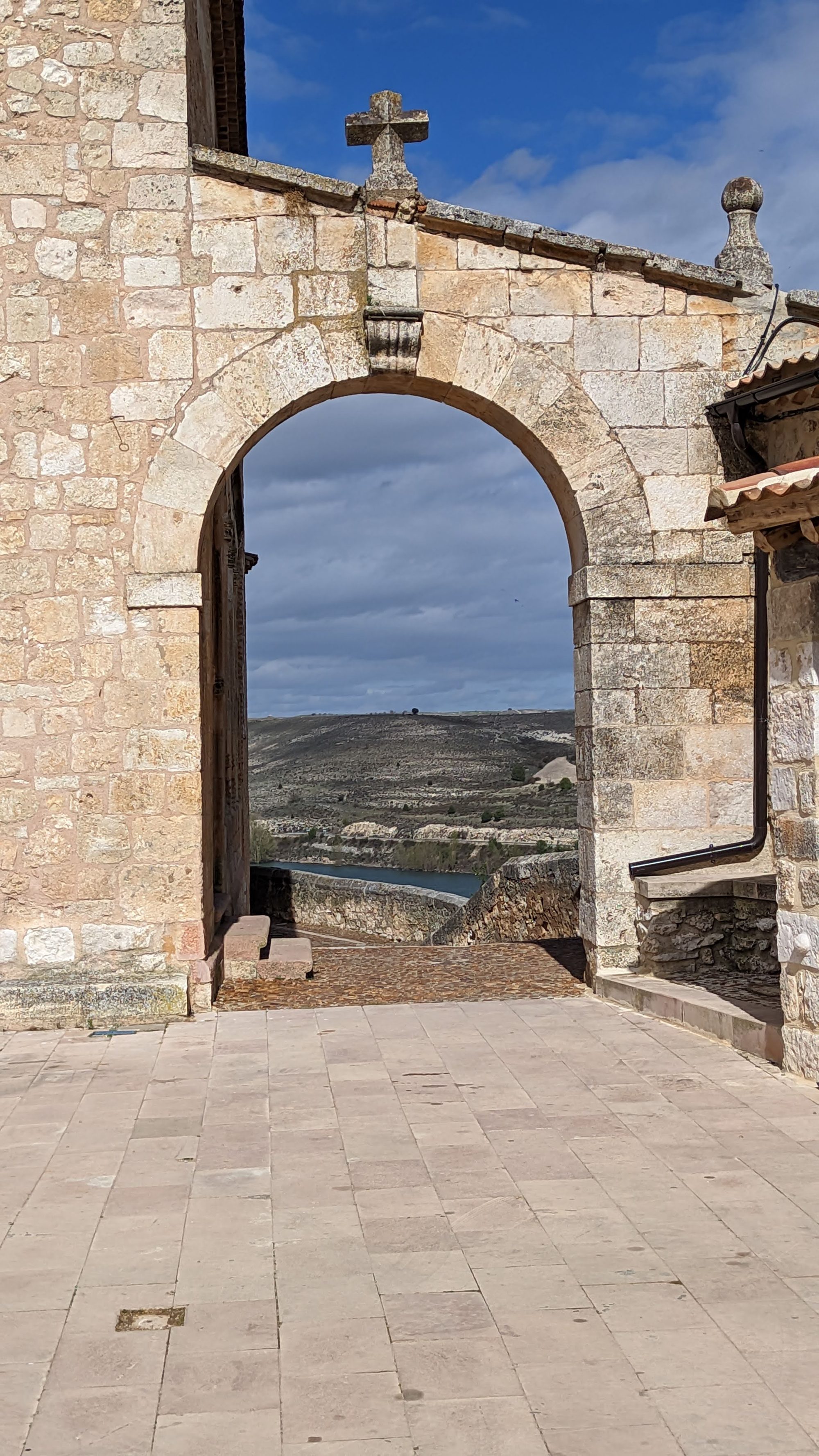 Arco de piedra con cruz tallada mostrando colinas y un embalse al fondo.