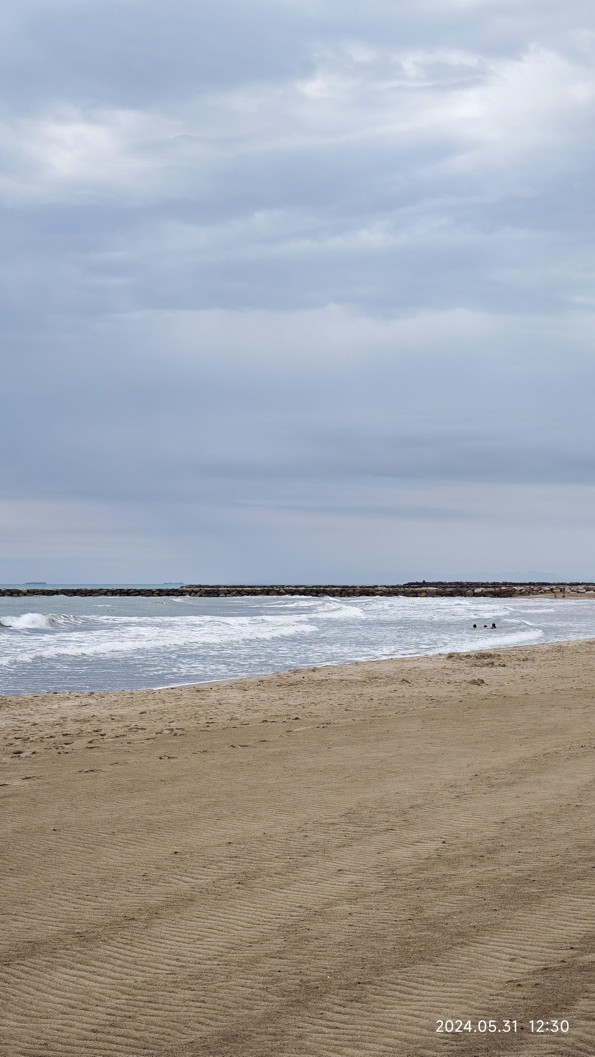 Playa de arena dorada con olas suaves y un espigón de piedras bajo cielo nublado.