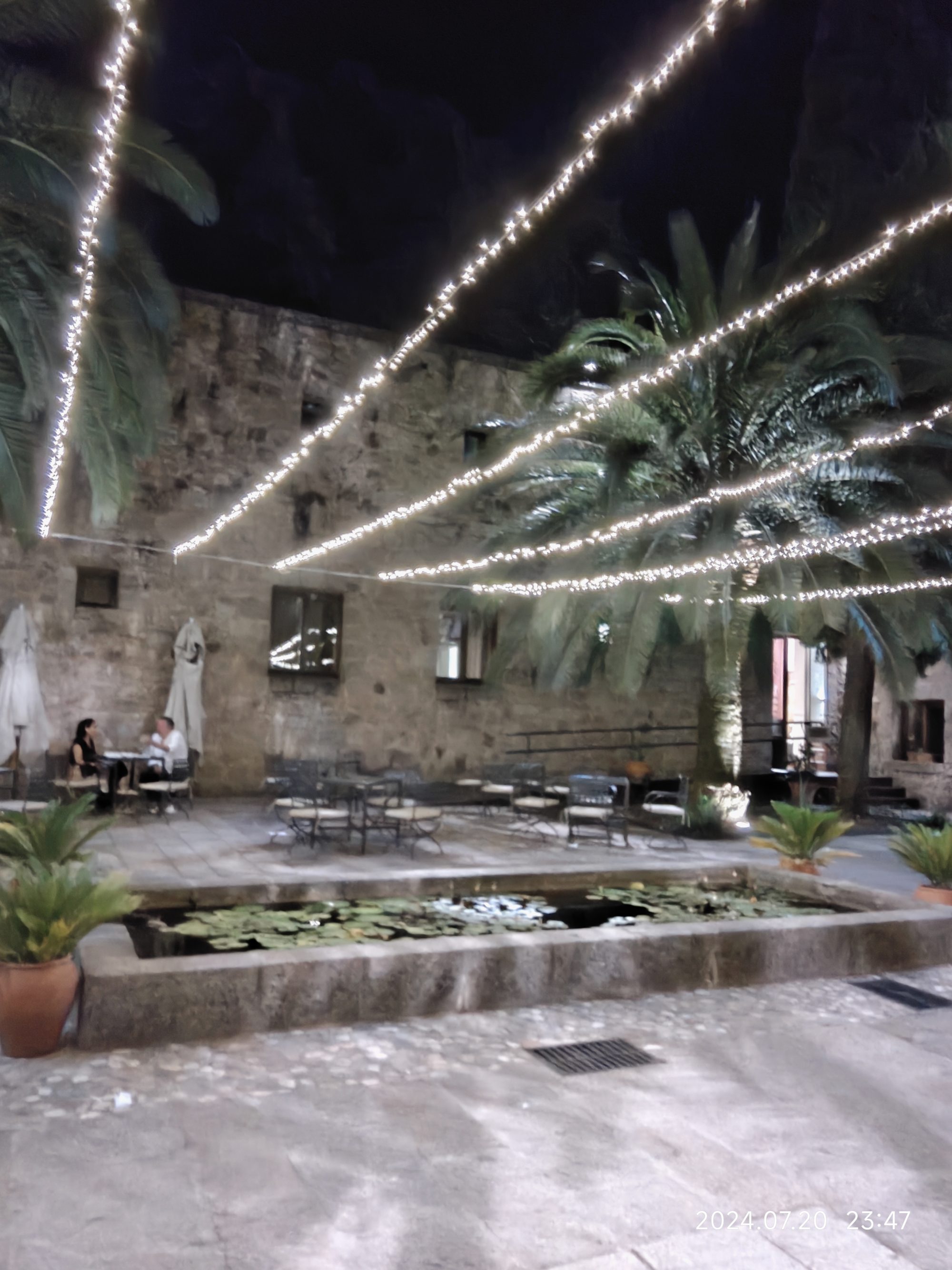 Patio de piedra antiguo iluminado con guirnaldas de luces, palmeras y estanque con nenúfares.