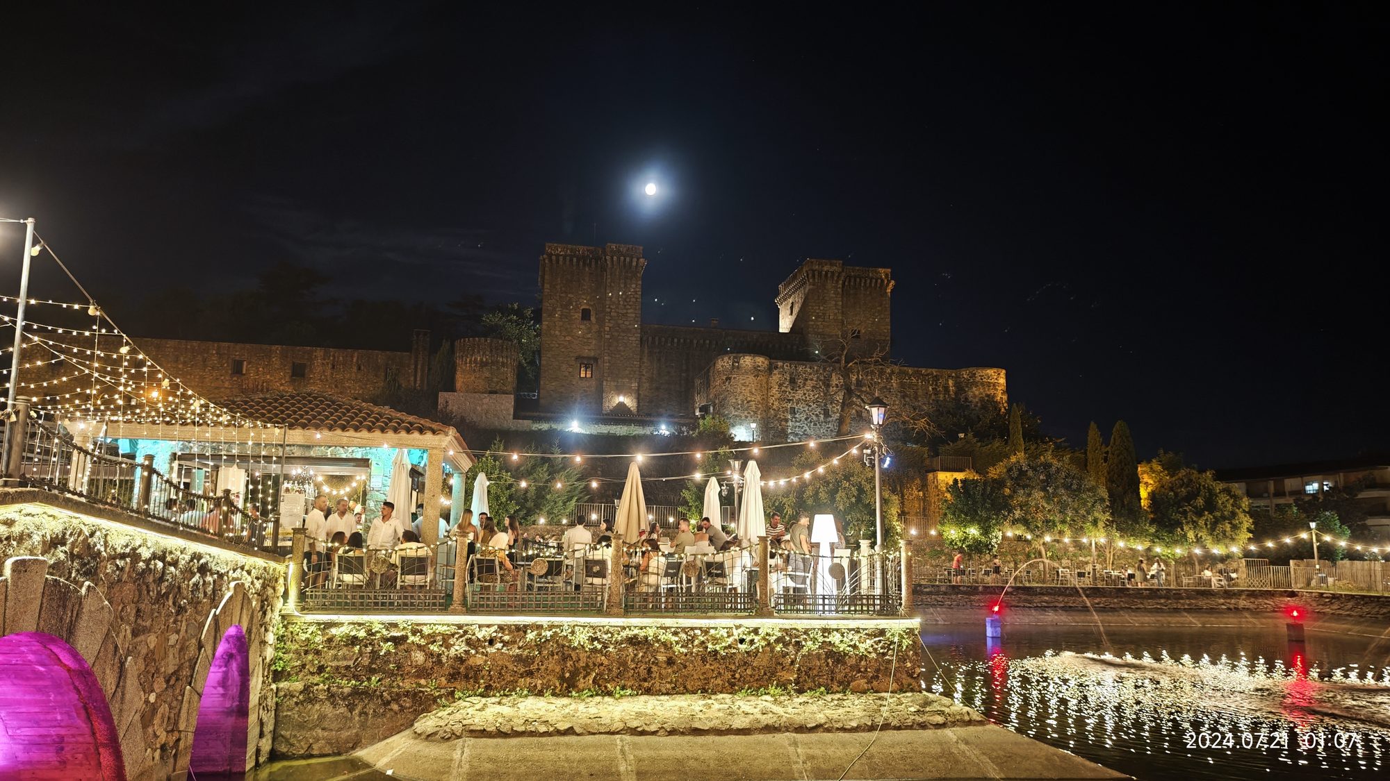 Castillo medieval iluminado de noche bajo la luna llena y terraza con luces festivas.