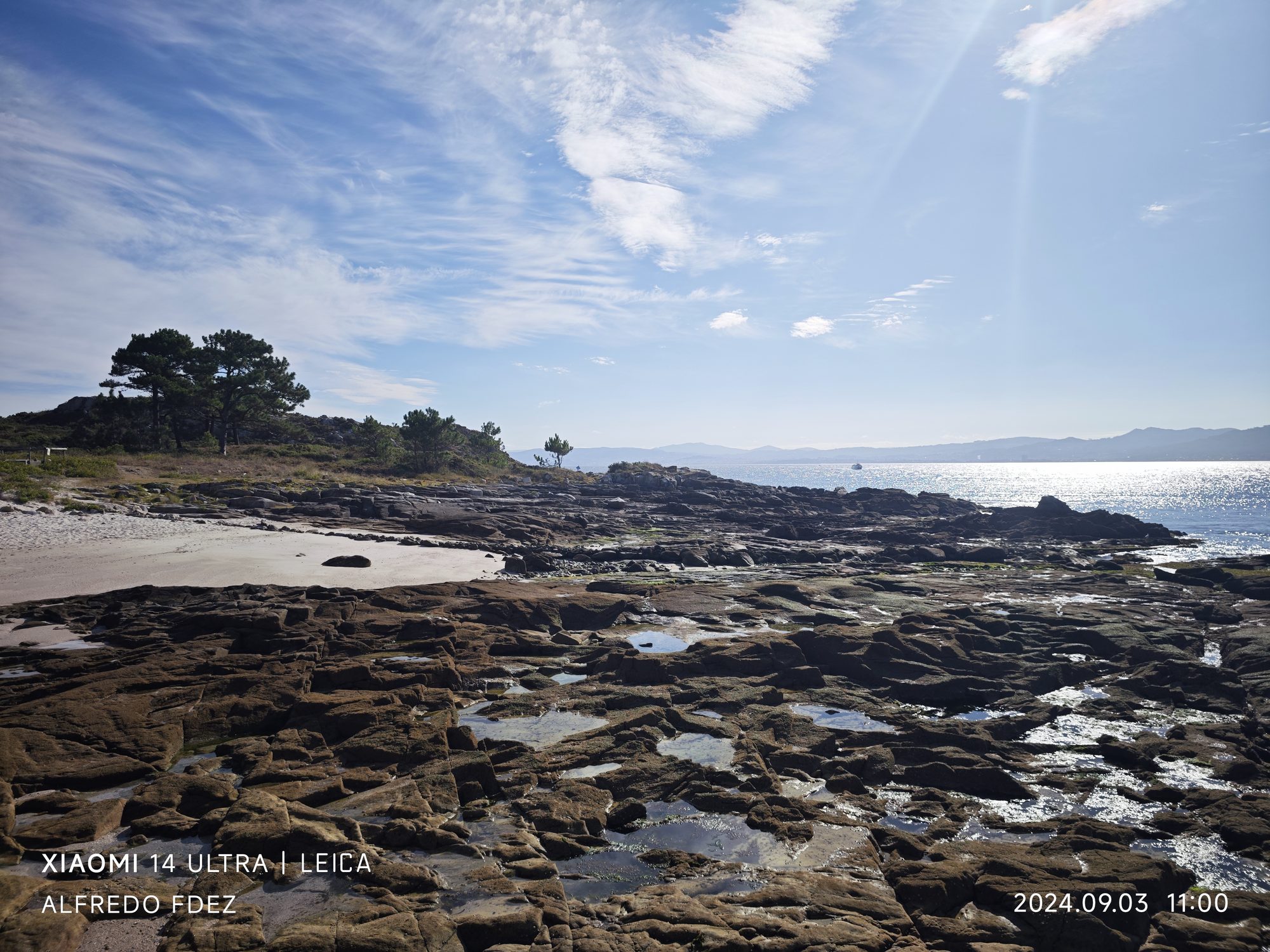 Costa rocosa y pequeña playa con pinos bajo el cielo soleado en Vigo.