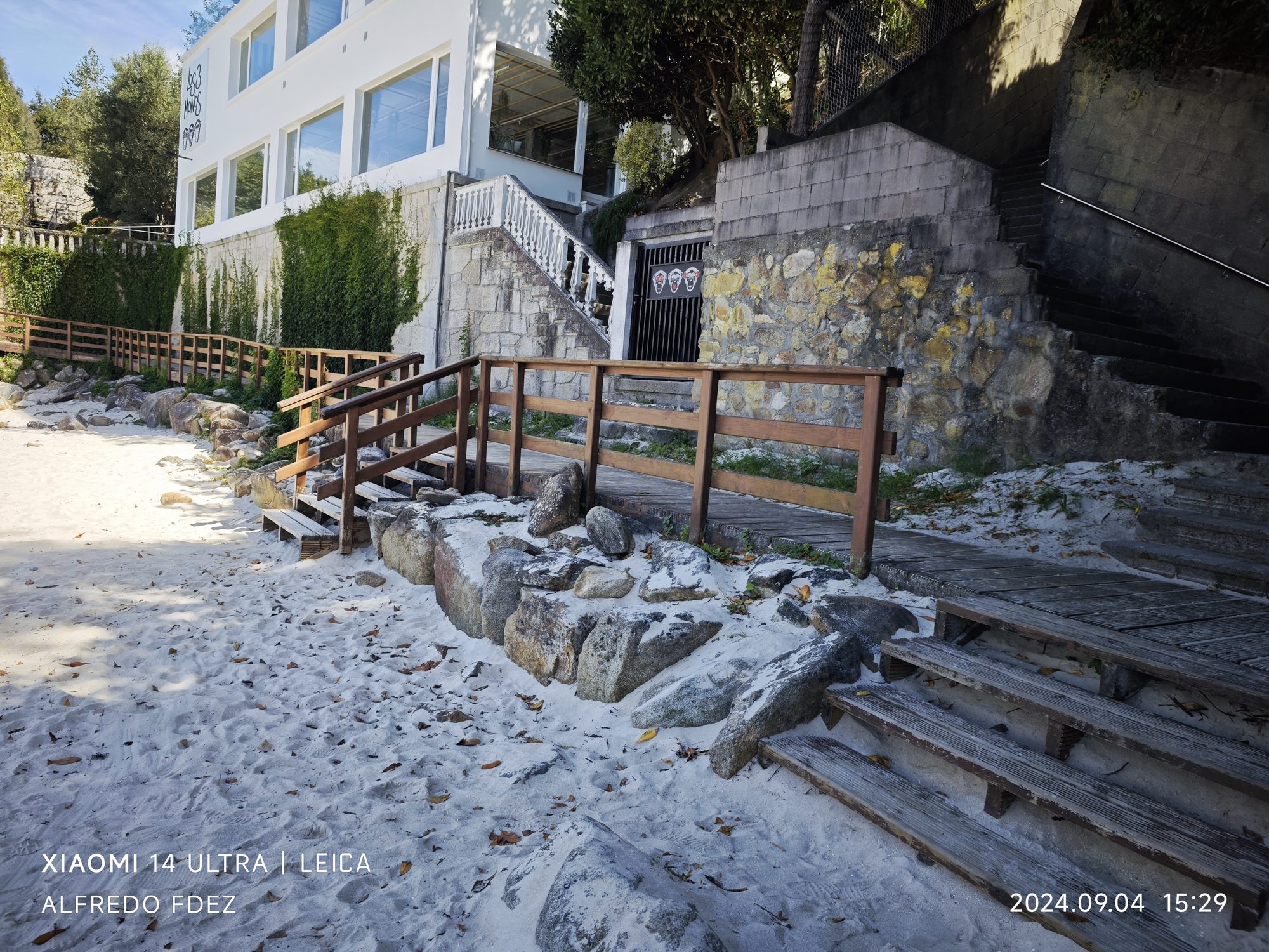 Acceso de madera a la playa con escaleras entre rocas y arena blanca.