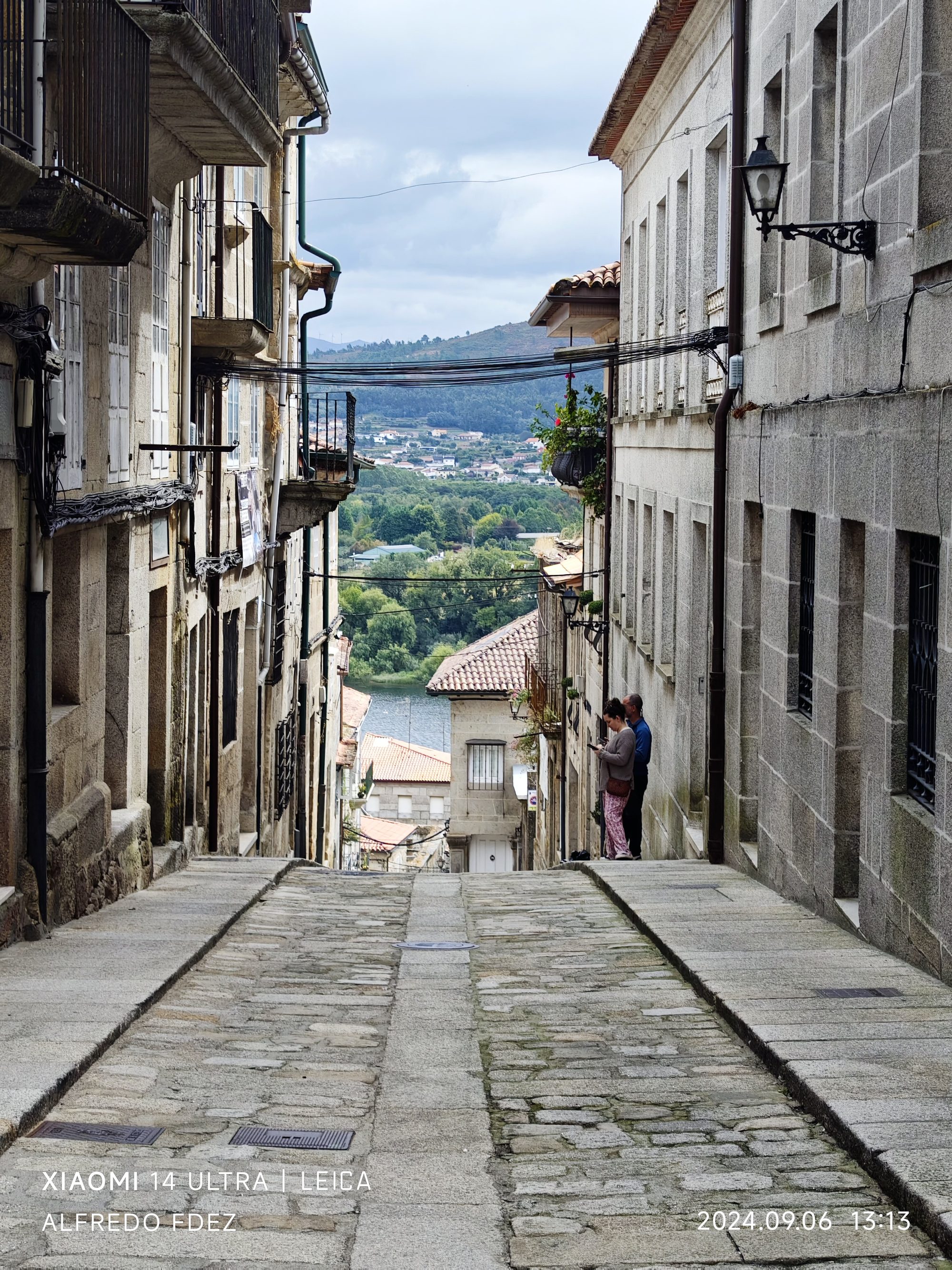 Calle empedrada empinada entre edificios de piedra en Tui con vistas al río y colinas.