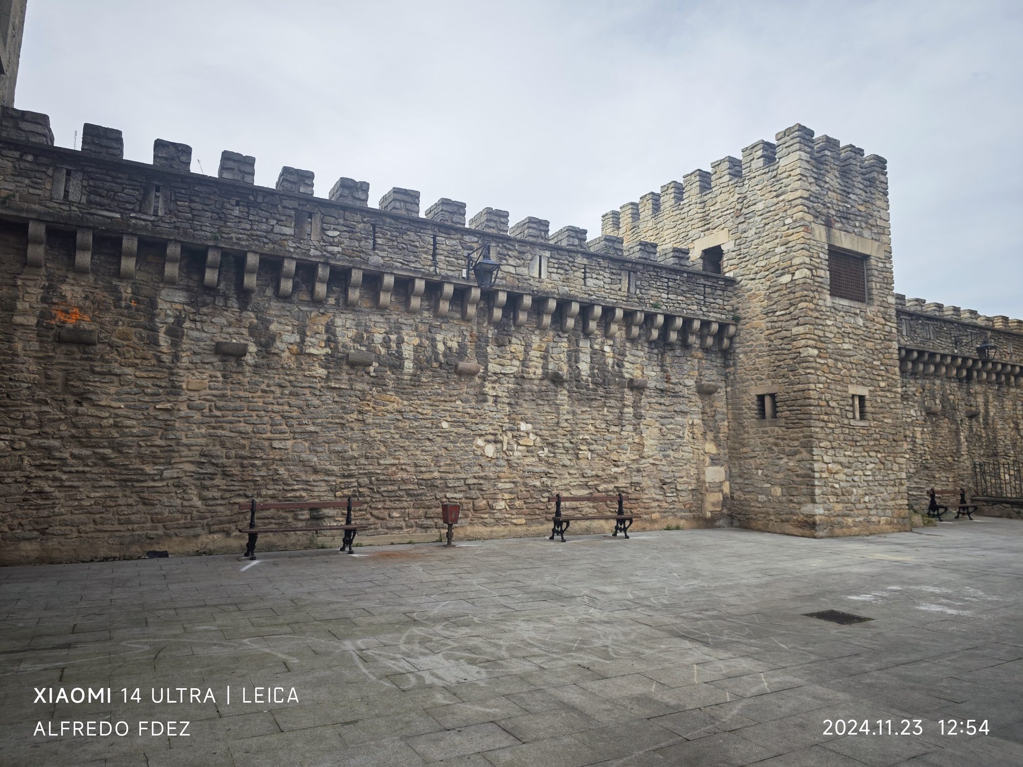 Muralla medieval de piedra con almenas y bancos de madera sobre una plaza pavimentada.