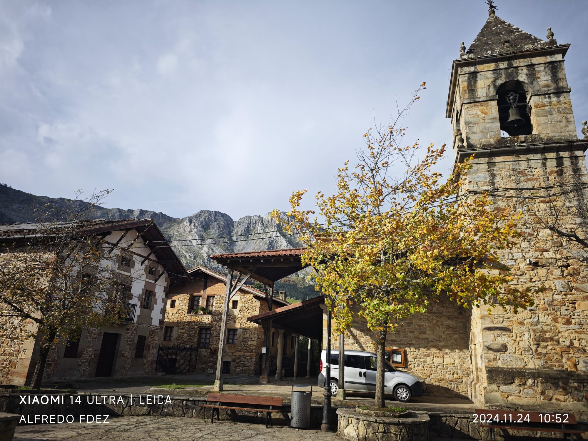 Torre de iglesia de piedra, casas tradicionales y montañas al fondo en Atxondo.