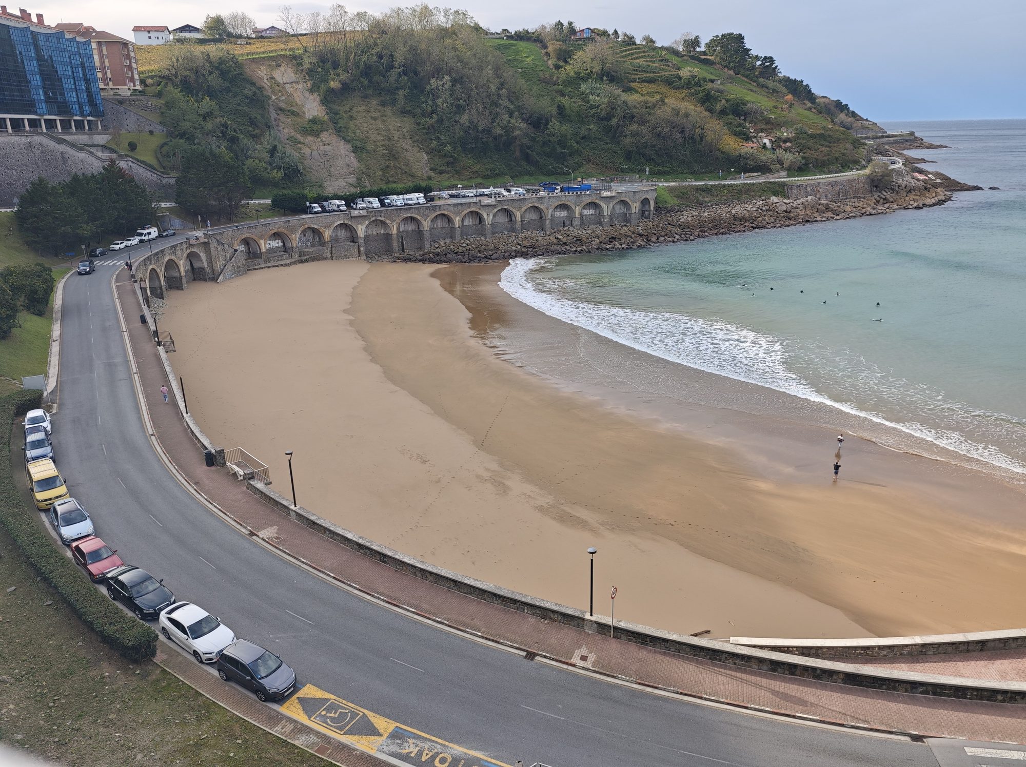 Playa semicircular de arena dorada junto a una carretera costera y colinas verdes en Guetaria.