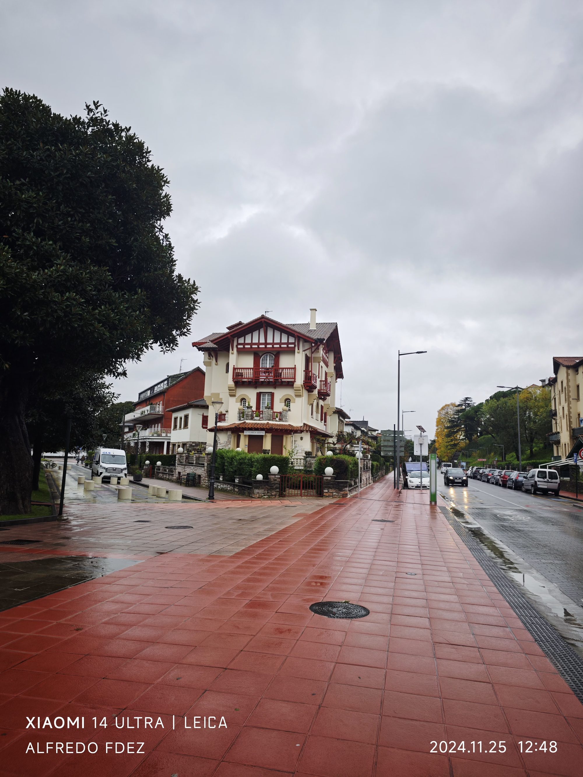 Casa tradicional vasca con entramado rojo junto a una acera mojada bajo cielo nublado.