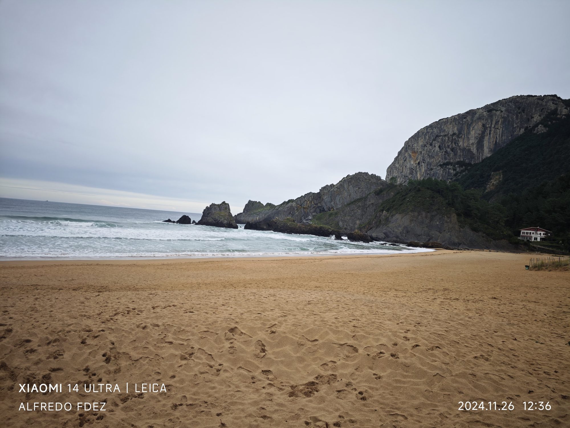 Playa de arena dorada con olas del mar y acantilados rocosos bajo cielo nublado.